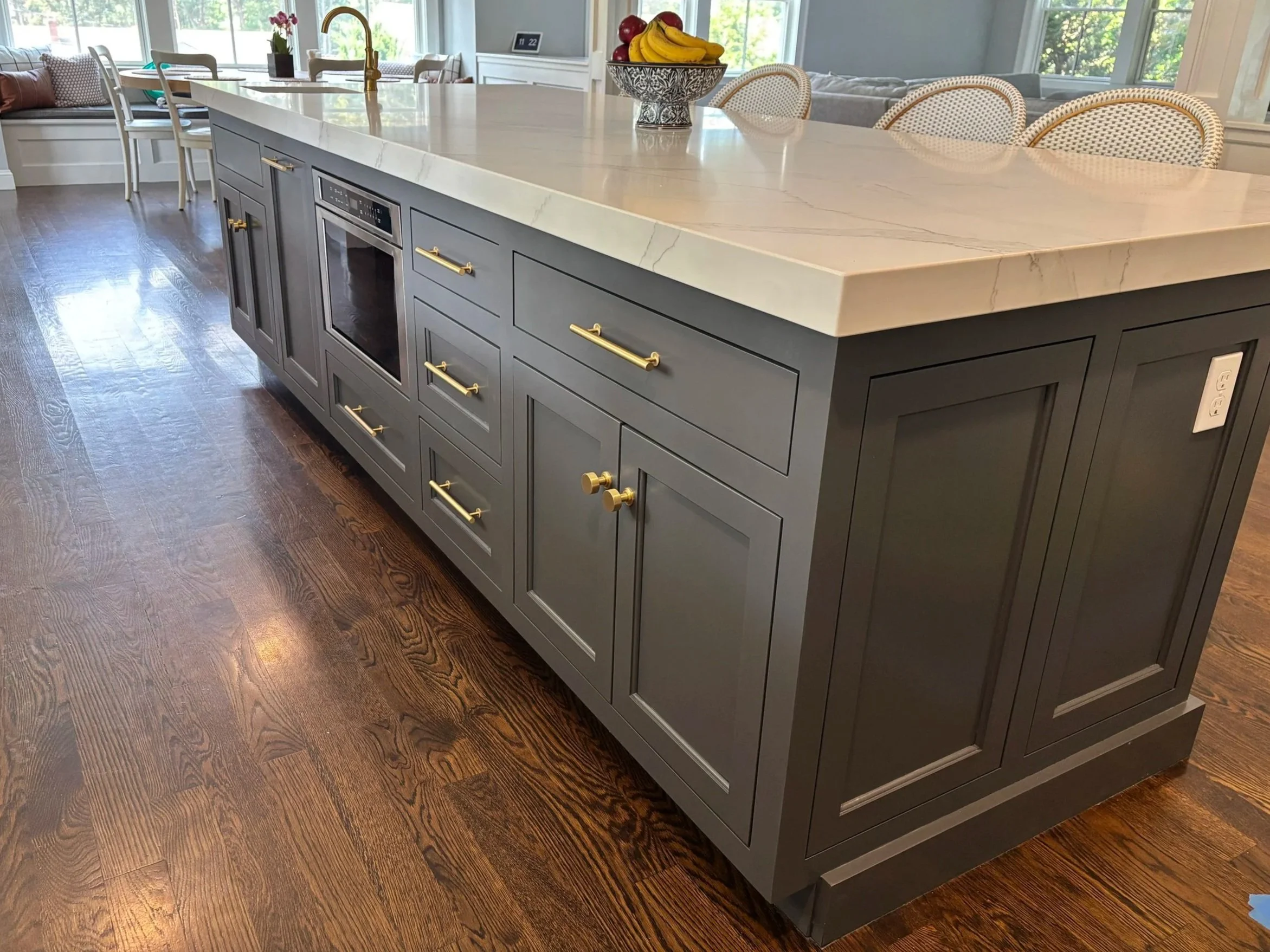 A kitchen island with a white marble countertop and dark gray cabinets with gold handles. There are chairs on one side, a decorative fruit bowl with bananas and red apples on top, and built-in appliances below. In the background, there is a dining area with a table, chairs, and large windows showing greenery outside.