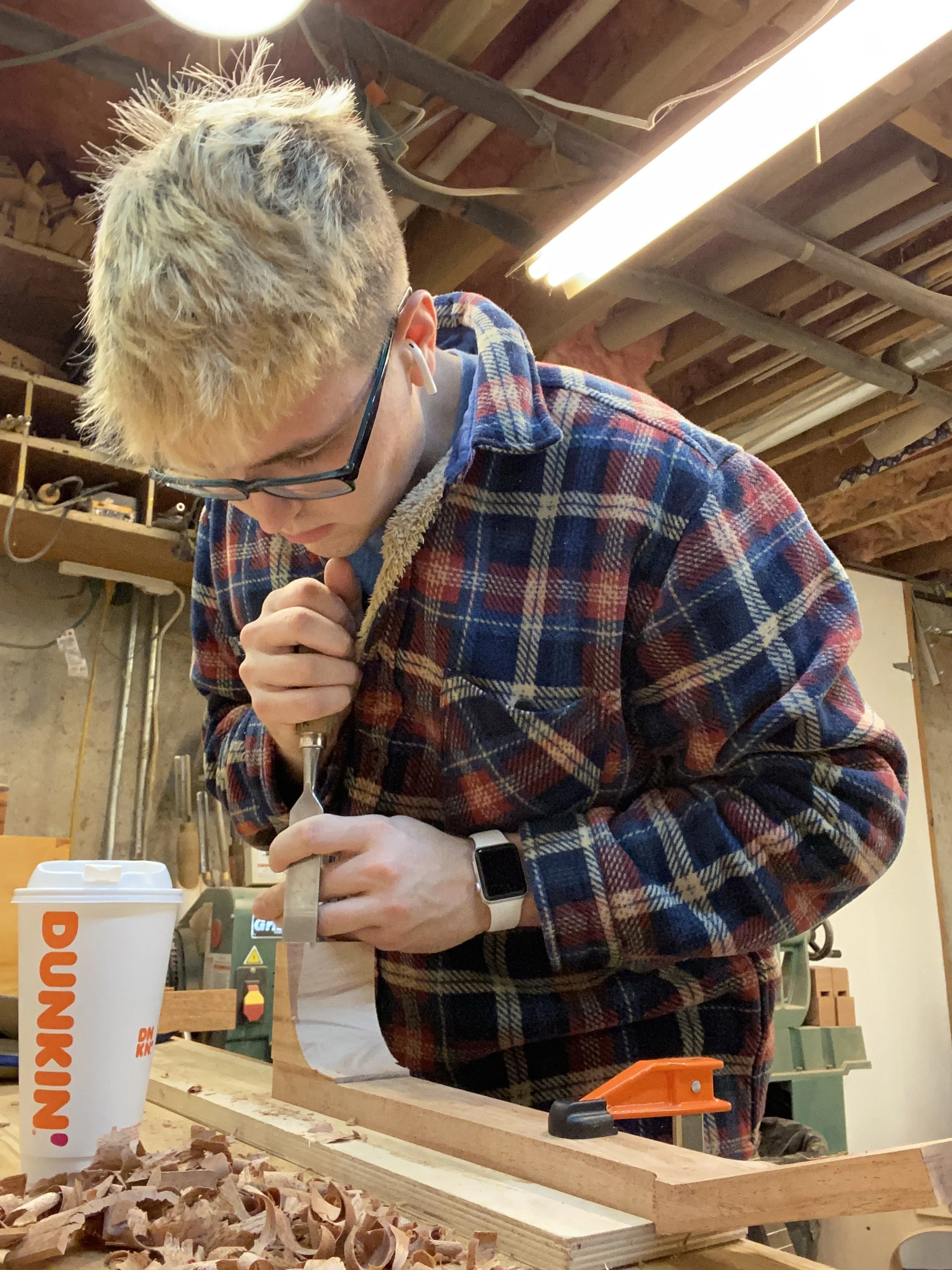 A person with blonde hair wearing glasses, a plaid shirt, and a watch, working in a woodworking shop, using a hand tool on a piece of wood. Wood shavings and work tools are visible in the foreground and background.