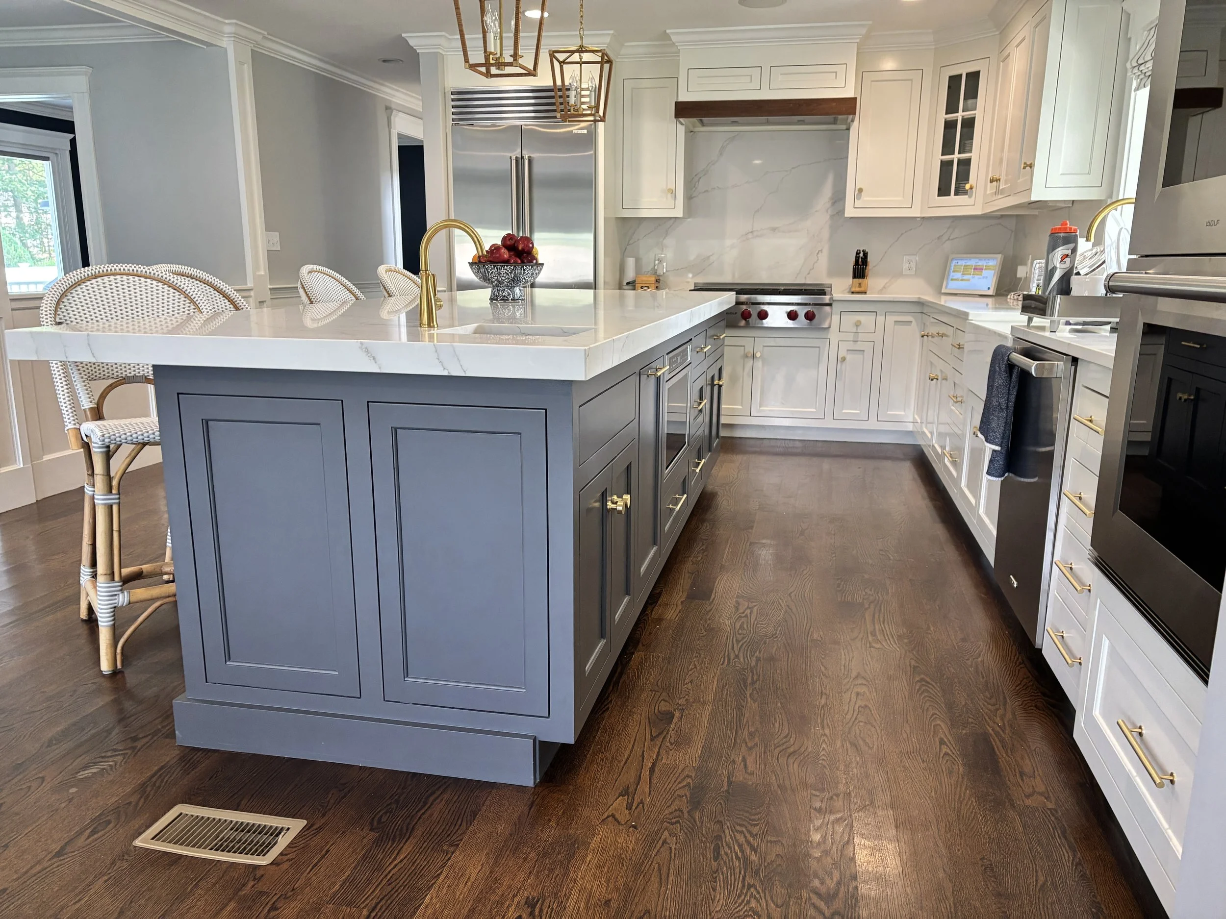 A modern kitchen with a large central island painted in dark gray with gold handles, topped with a white marble countertop. There are four wicker chairs with white cushions on one side. The kitchen features white cabinets with gold hardware, a stainless steel refrigerator, a stove with red knobs, and various kitchen gadgets. A bowl of red apples and a gold faucet are on the island. The floor is wood, and there is a vent on the floor near the island.
