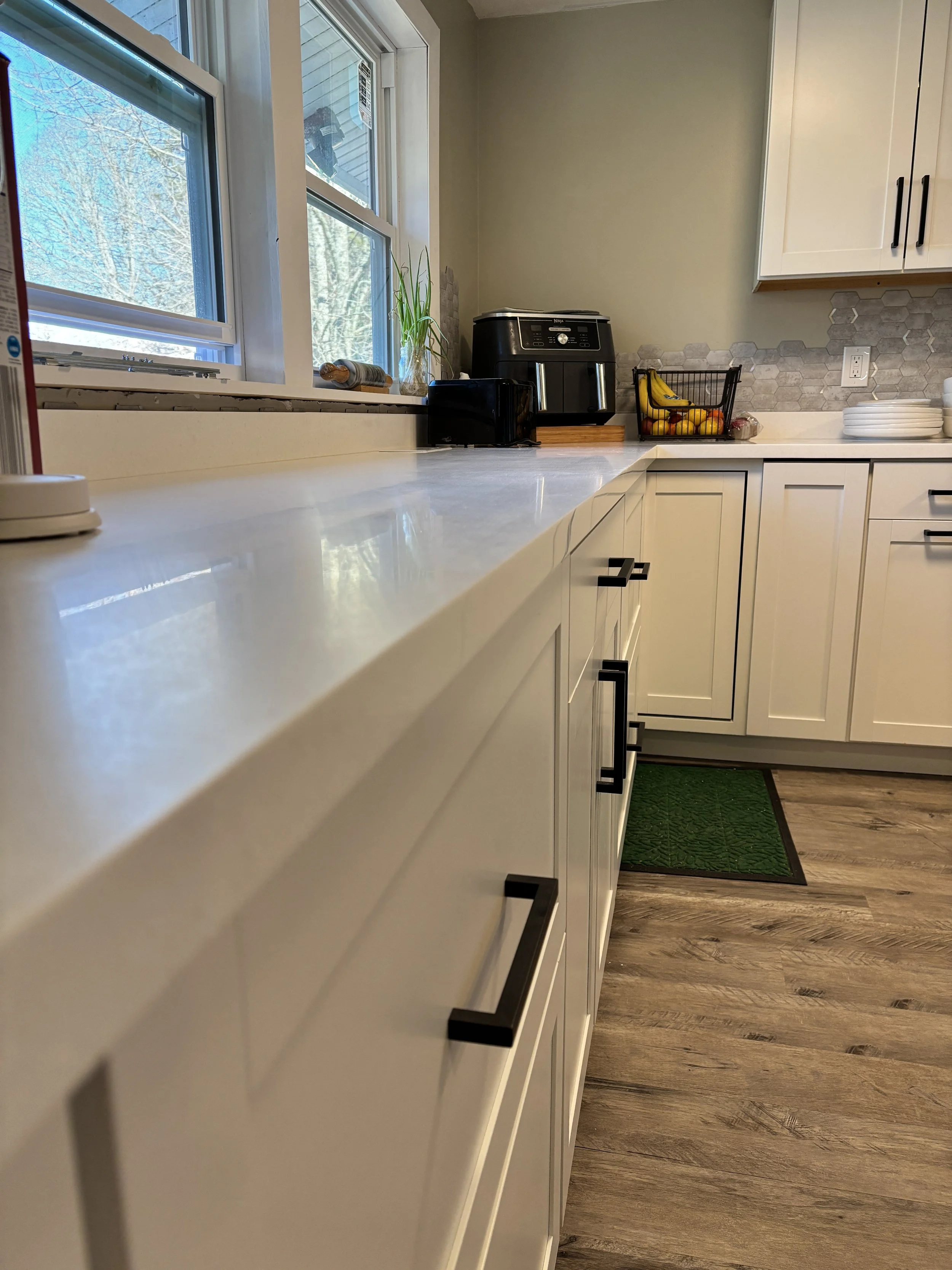 A kitchen countertop with a white surface, featuring a black toaster, a black air fryer, a small plant in a glass jar, a basket of bananas and fruits, white plates stacked, and a green rug on a wooden floor with wallpapered and cabinet walls.