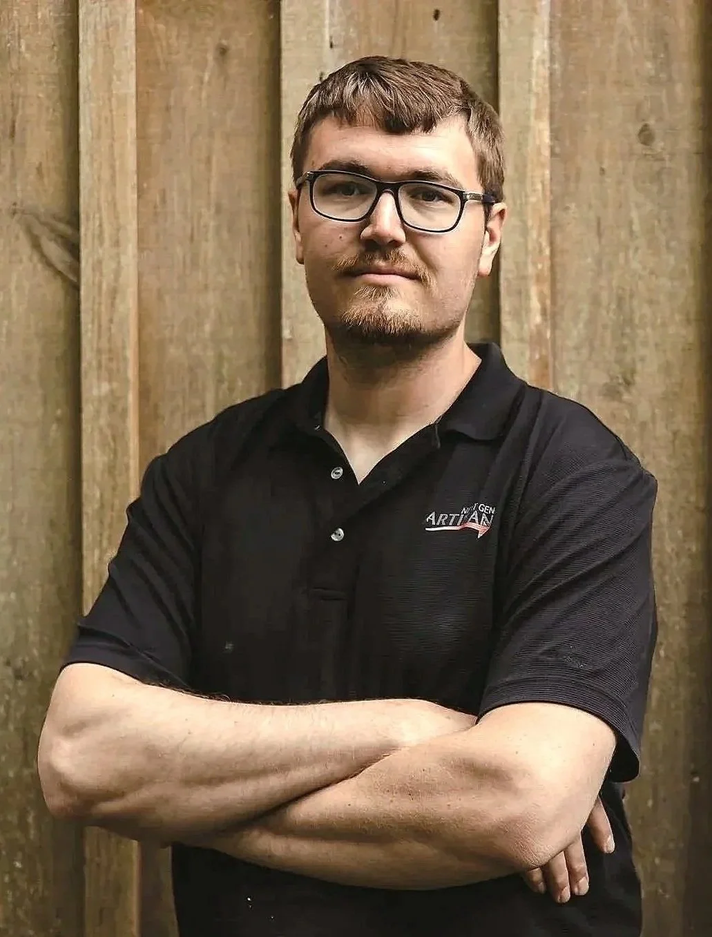 A man with glasses, facial hair, and short brown hair posing with crossed arms in front of a wooden background. He is wearing a black polo shirt with a logo on the left chest.