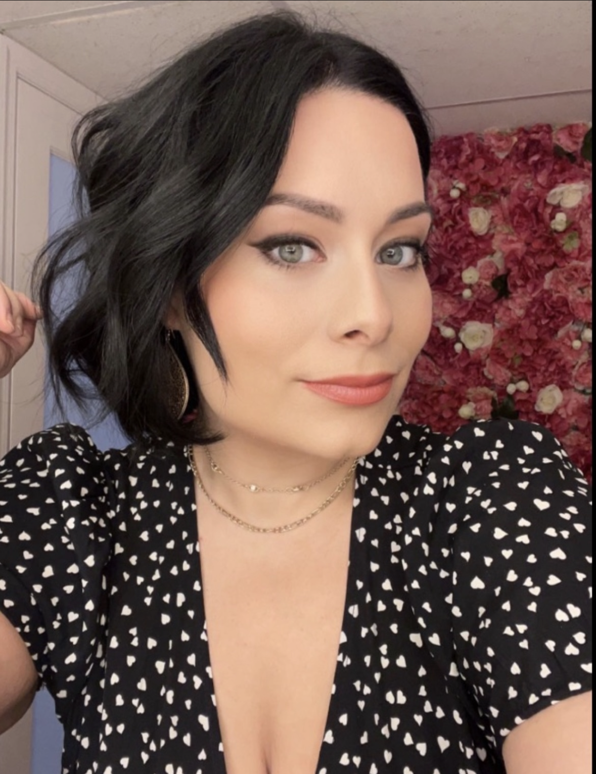 A woman with short black hair, light skin, and gray eyes takes a selfie indoors in front of a pink and white flower-covered wall. She wears a black dress with white heart patterns, gold earrings, and layered gold necklaces, with makeup including pink lipstick and eyeliner.