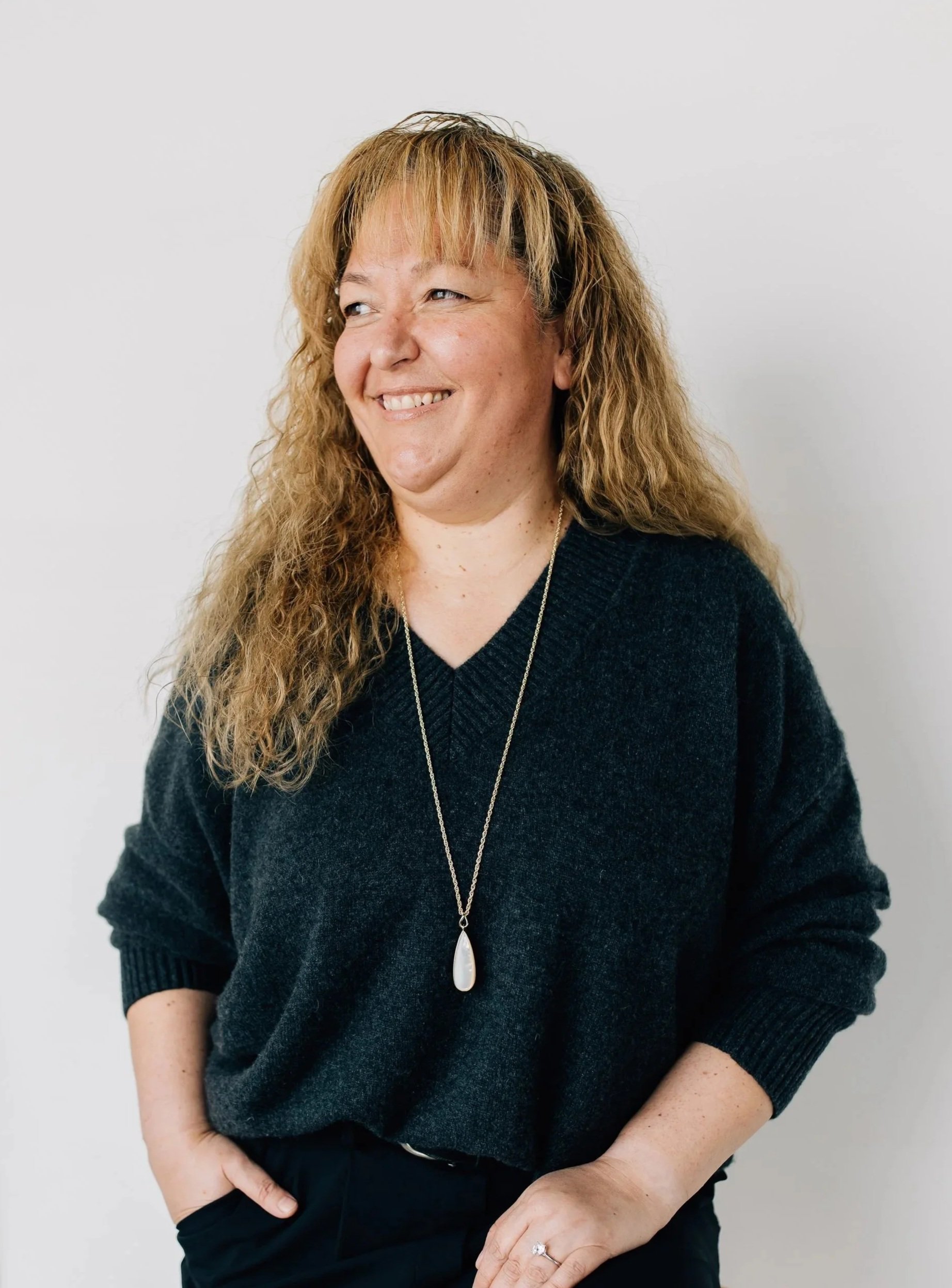 Liliana Baylon, a Latina woman with long golden brown curly hair, is wearing a dark grey jumper, looking to her right with a silver necklace with a tear drop shaped pendant.