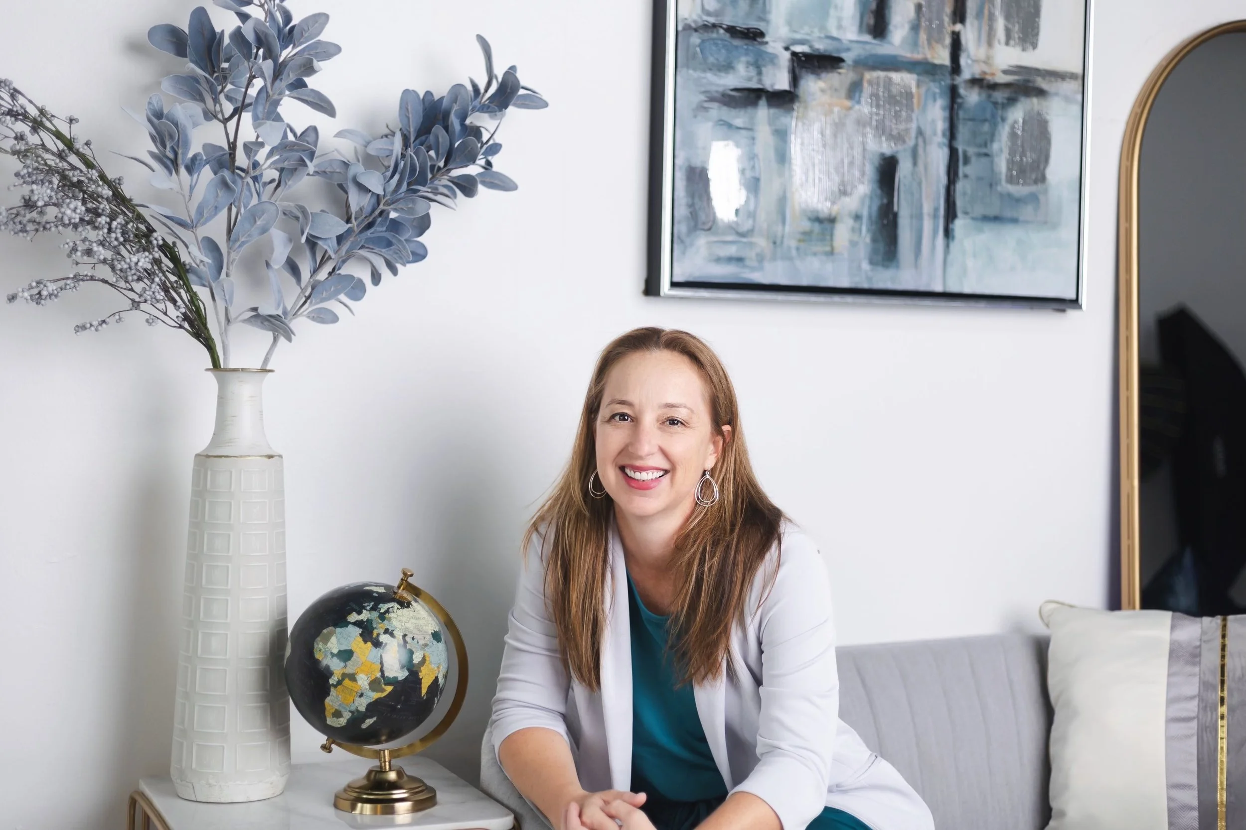Photo of Jen Taylor smiling next to a vase of flowers and sitting on a sofa. She has long straight brown hair and is wearing a white suit jacket.