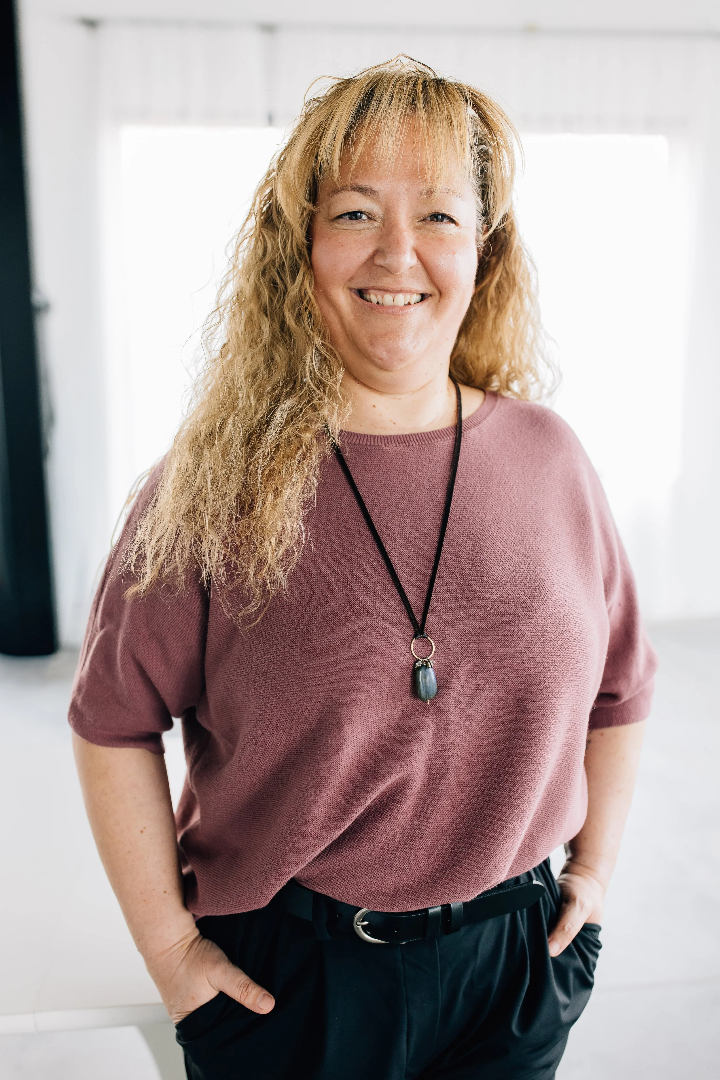 A smiling woman with long, curly gray hair sitting on a beige couch, wearing a gray top, black pants, and a necklace with a large dark heart-shaped pendant.