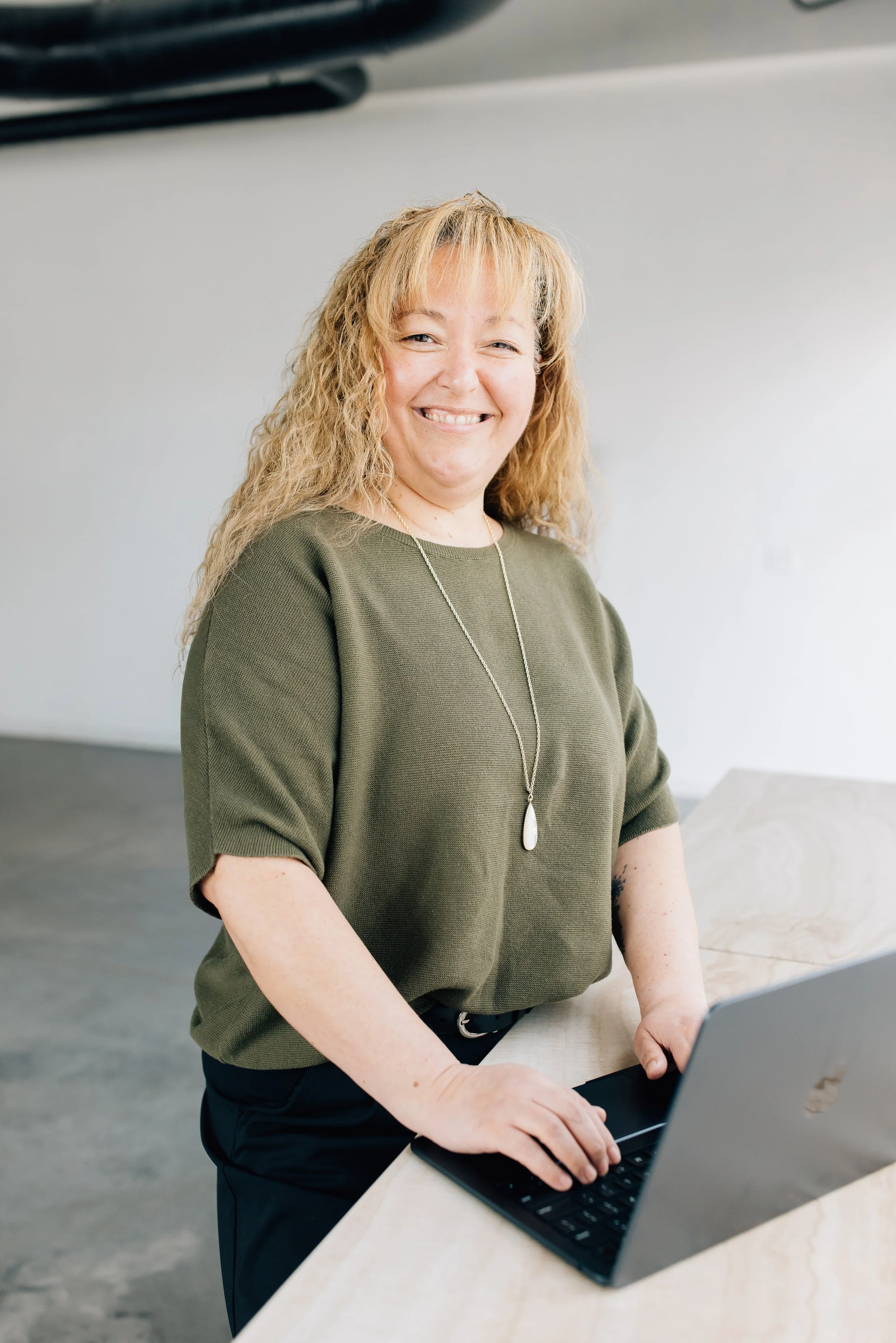 Liliana Baylon, a Latina woman with long golden brown curly hair, is wearing a green top and silver necklace standing at a desk with her hands typing on a laptop.
