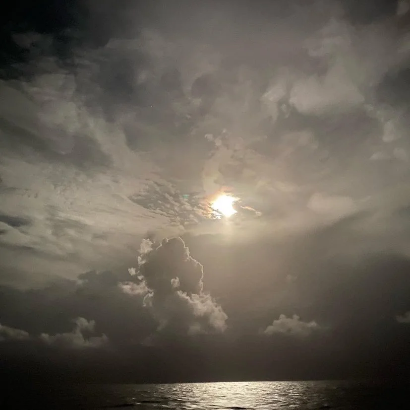 Nighttime ocean scene with a cloudy sky and a bright moon shining through the clouds.