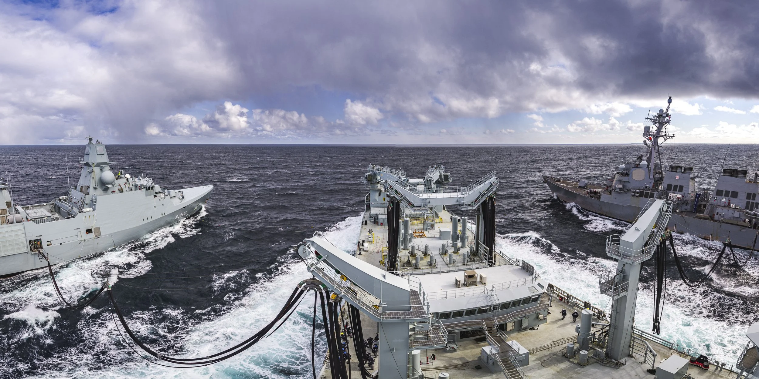 View from the deck of a navy ship with two other ships nearby in rough ocean waters under a cloudy sky.
