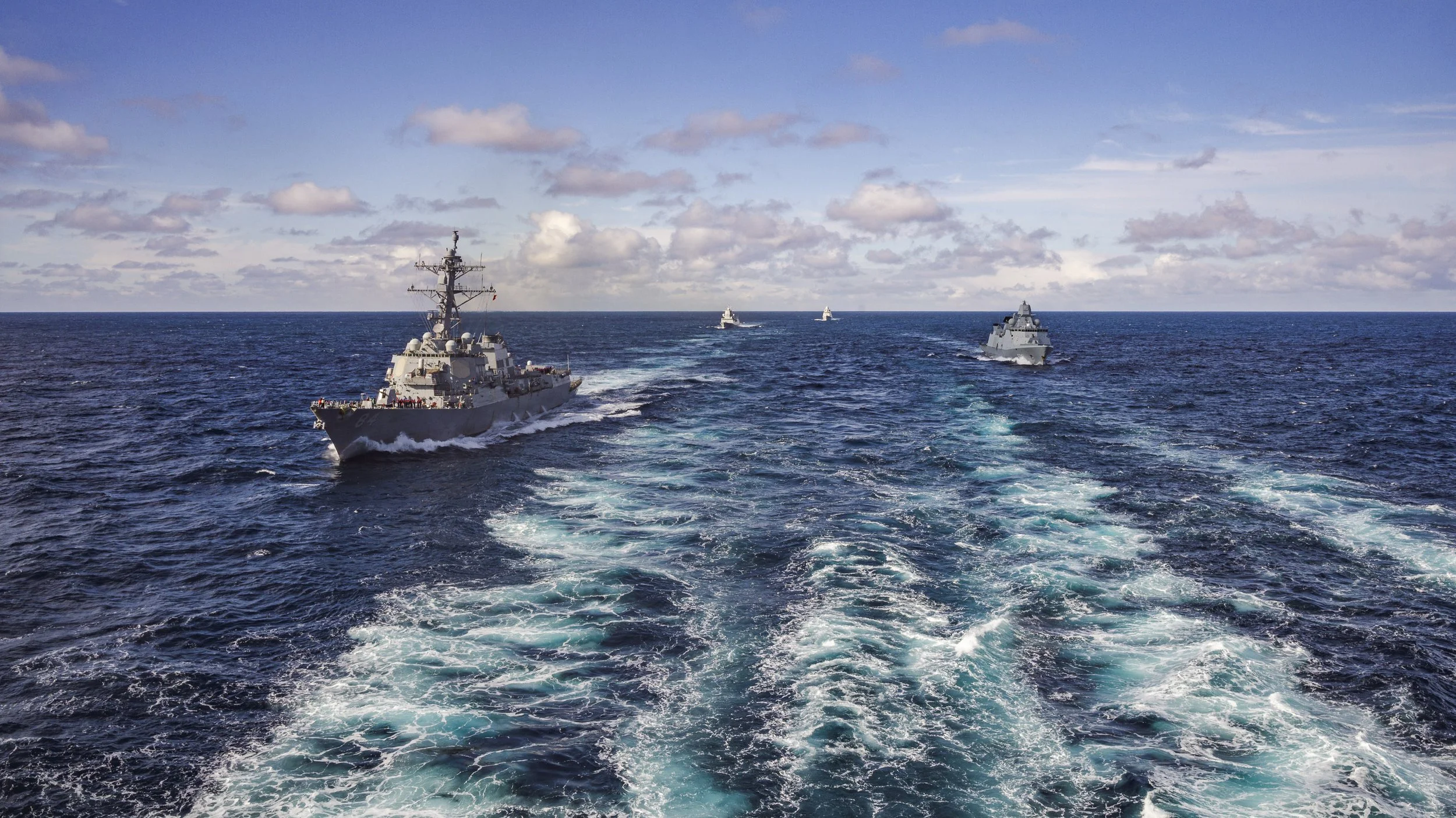 Four navy ships sailing on the ocean, leaving white splashes behind as they move forward under a partly cloudy sky.