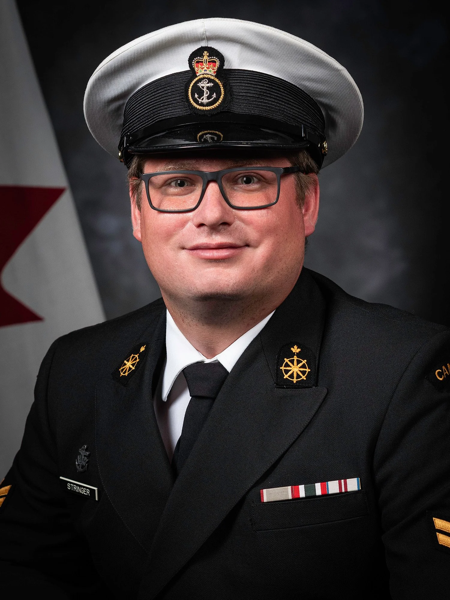 Portrait of a man in a formal military uniform, including a captain's hat, glasses, and various medals, with a flag in the background.