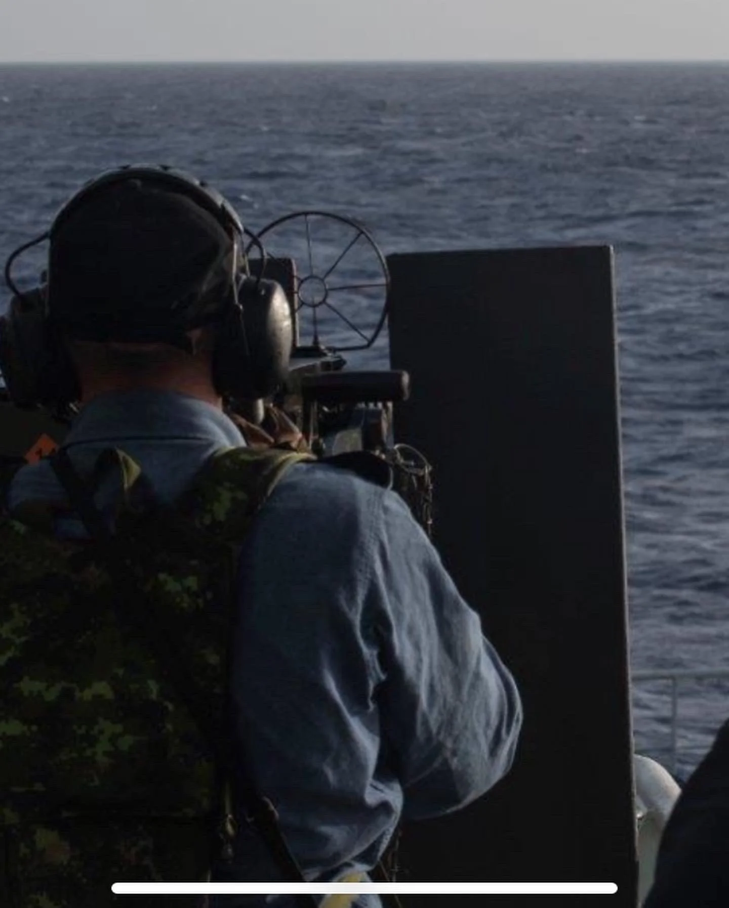 A person in military or tactical gear, wearing a helmet and headphones, operating a weapon or equipment on a boat or ship, looking out over the ocean during daylight.
