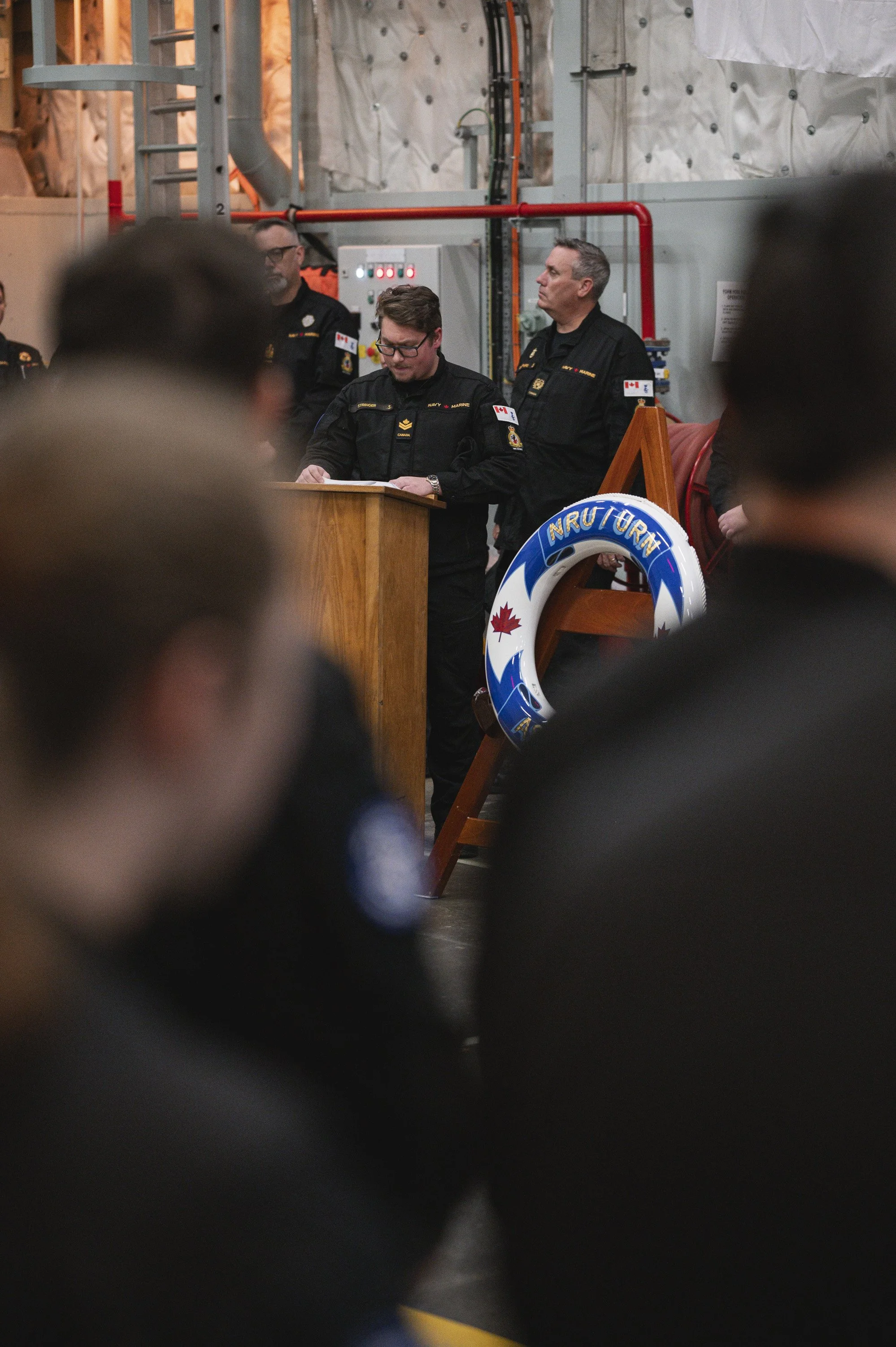 Firefighters in black uniforms standing behind a wooden podium during a ceremony, with a life ring labeled 'RESCUE' and Canadian flags visible.
