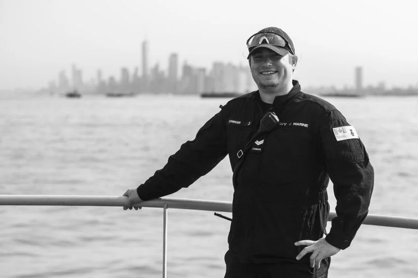 A man dressed in a dark uniform with Marine insignia, wearing sunglasses, standing on a boat with a city skyline and water in the background.