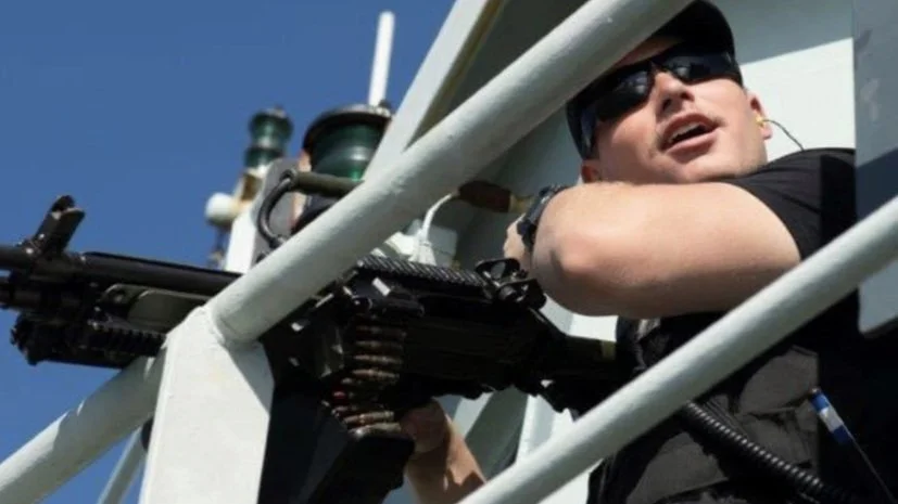 A person wearing sunglasses and a black shirt operating a mounted machine gun on a white metal structure under a clear blue sky.