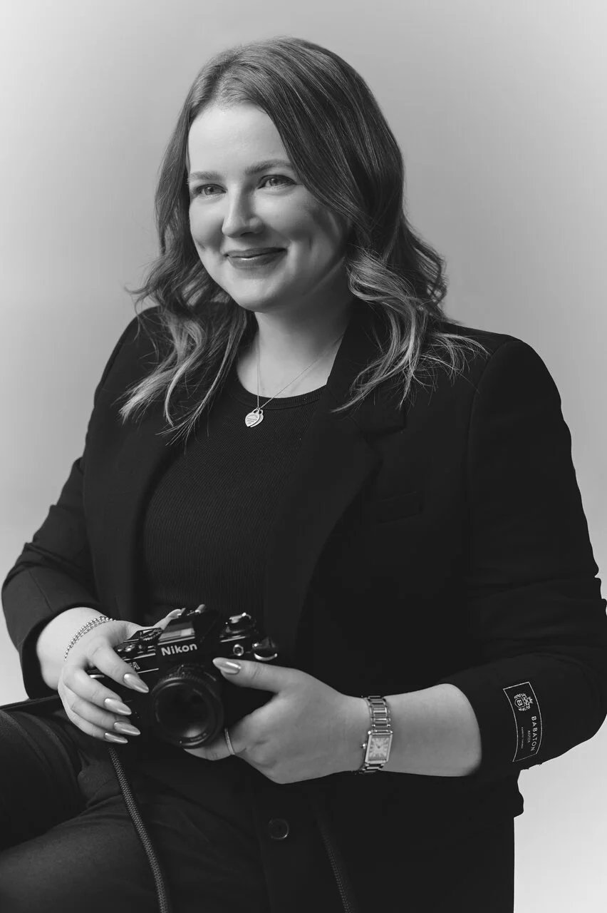 Black and white portrait of a woman holding a Nikon camera, smiling slightly, wearing a blazer, a necklace, and a watch, with wavy hair.