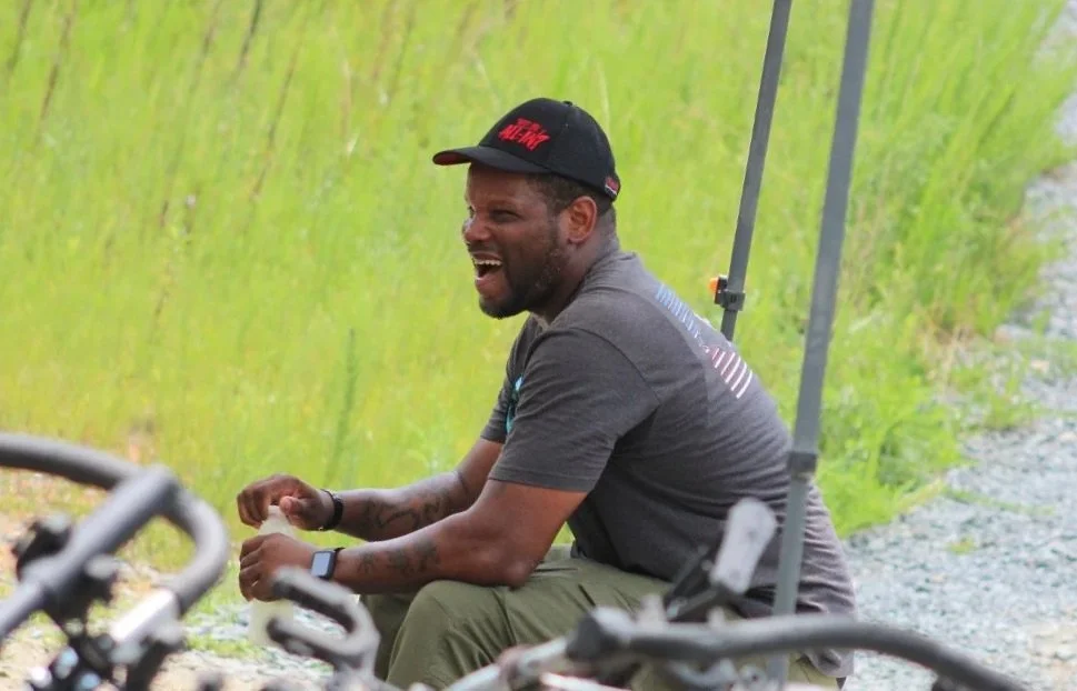 A man sitting outdoors on a gravel path with a green grassy background, smiling and laughing, wearing a black cap and a dark t-shirt.