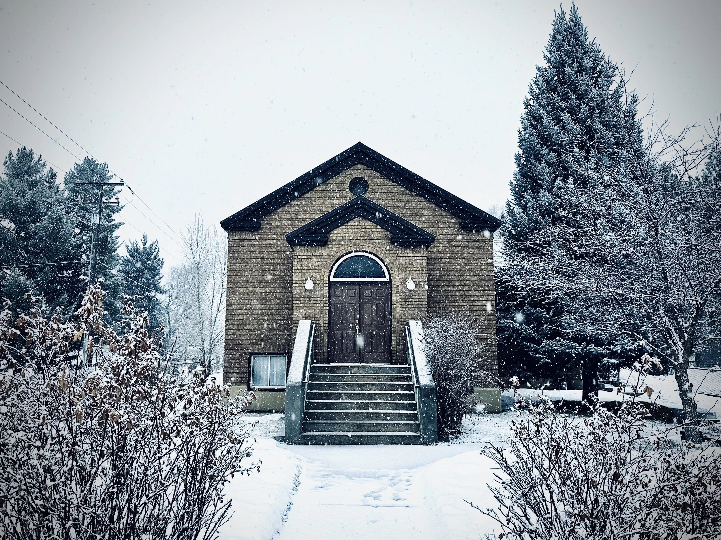 A brick church surrounded by snow-covered trees and bushes, with a set of stairs leading to a wooden door, and snow falling gently.
