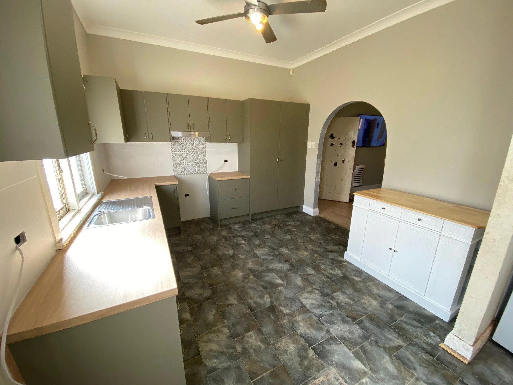 A new kitchen installed with light wood countertops, and a gray tile backsplash. There is a window above the sink, a ceiling fan with light, and a white sideboard with a light wood top. The floor is covered in dark gray tiles.