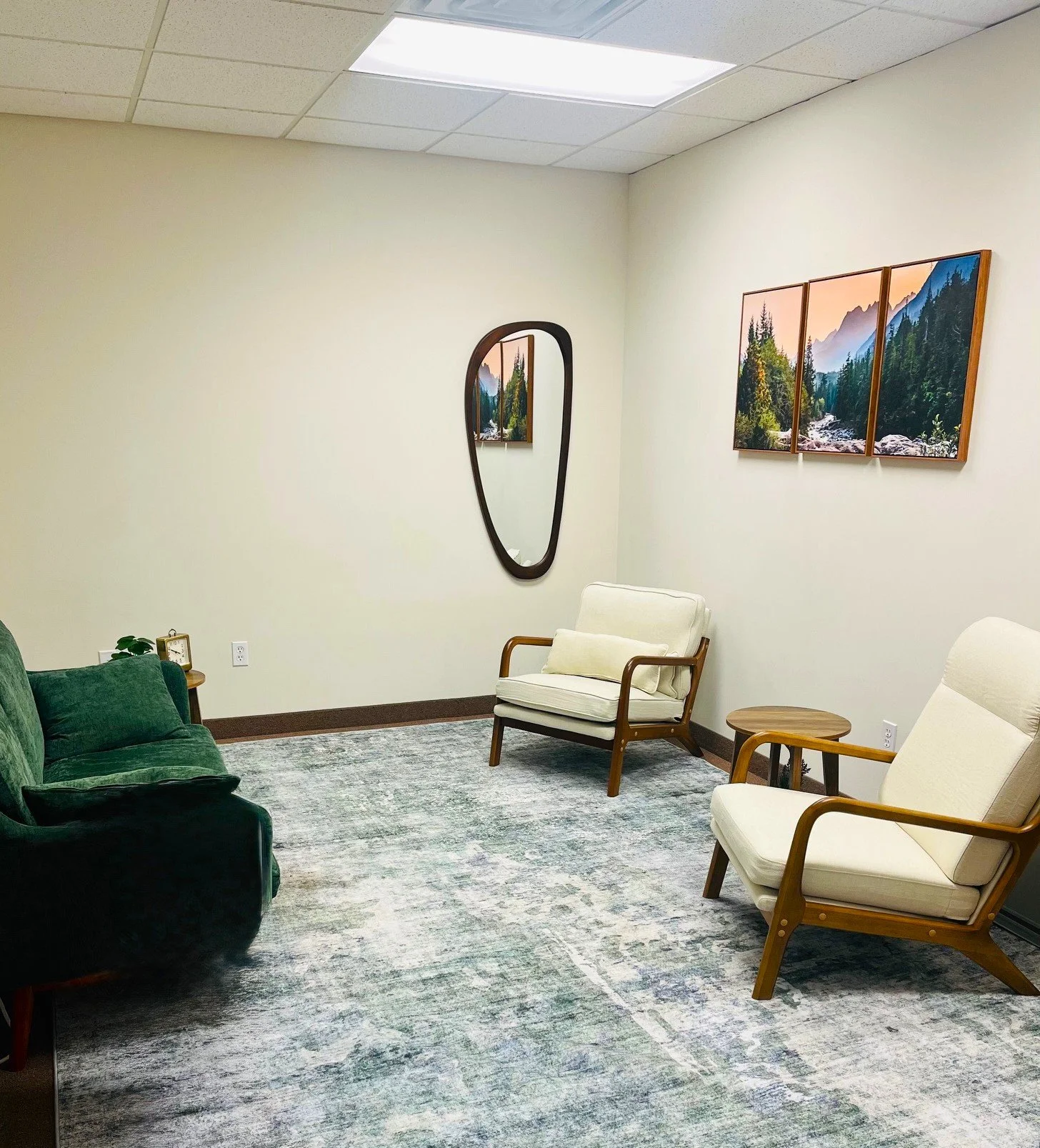 A waiting room with cream-colored walls, a large landscape painting of mountains and trees, and a mirror on the wall. There are chairs and a sofa visible, with a small side table holding a clock and a plant.