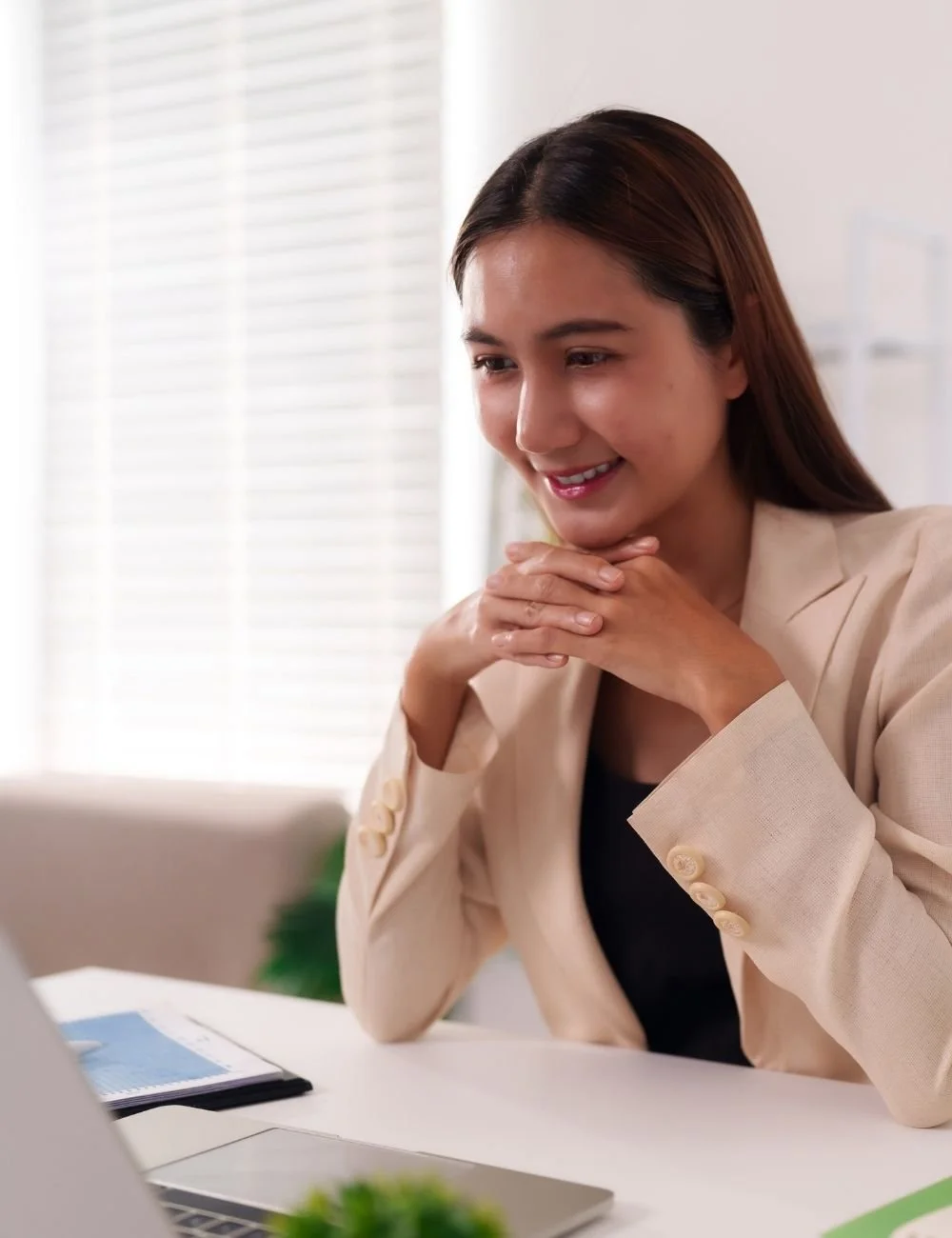A woman in business attire smiling and sitting at a desk with a laptop in front of her.