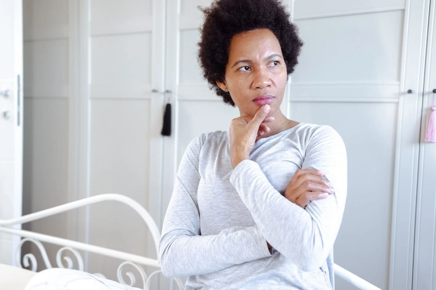A woman with short curly hair, wearing a light gray long-sleeve shirt, standing in a room with white cabinets and a white bed frame, appears to be deep in thought.