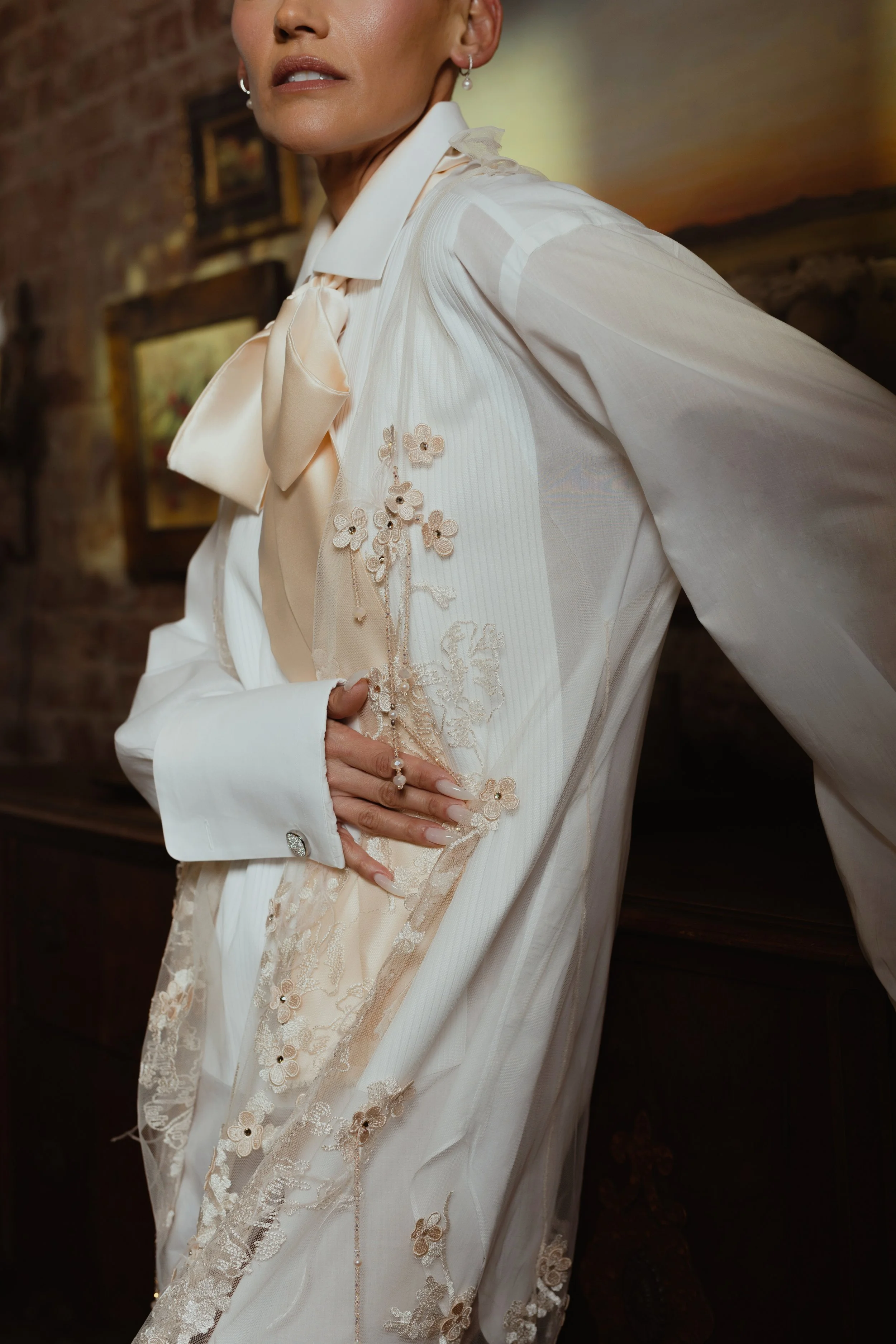 Close-up of a woman wearing an elegant cream-colored blouse with floral embroidery and lace detailing, standing indoors with a brick wall and paintings in the background.