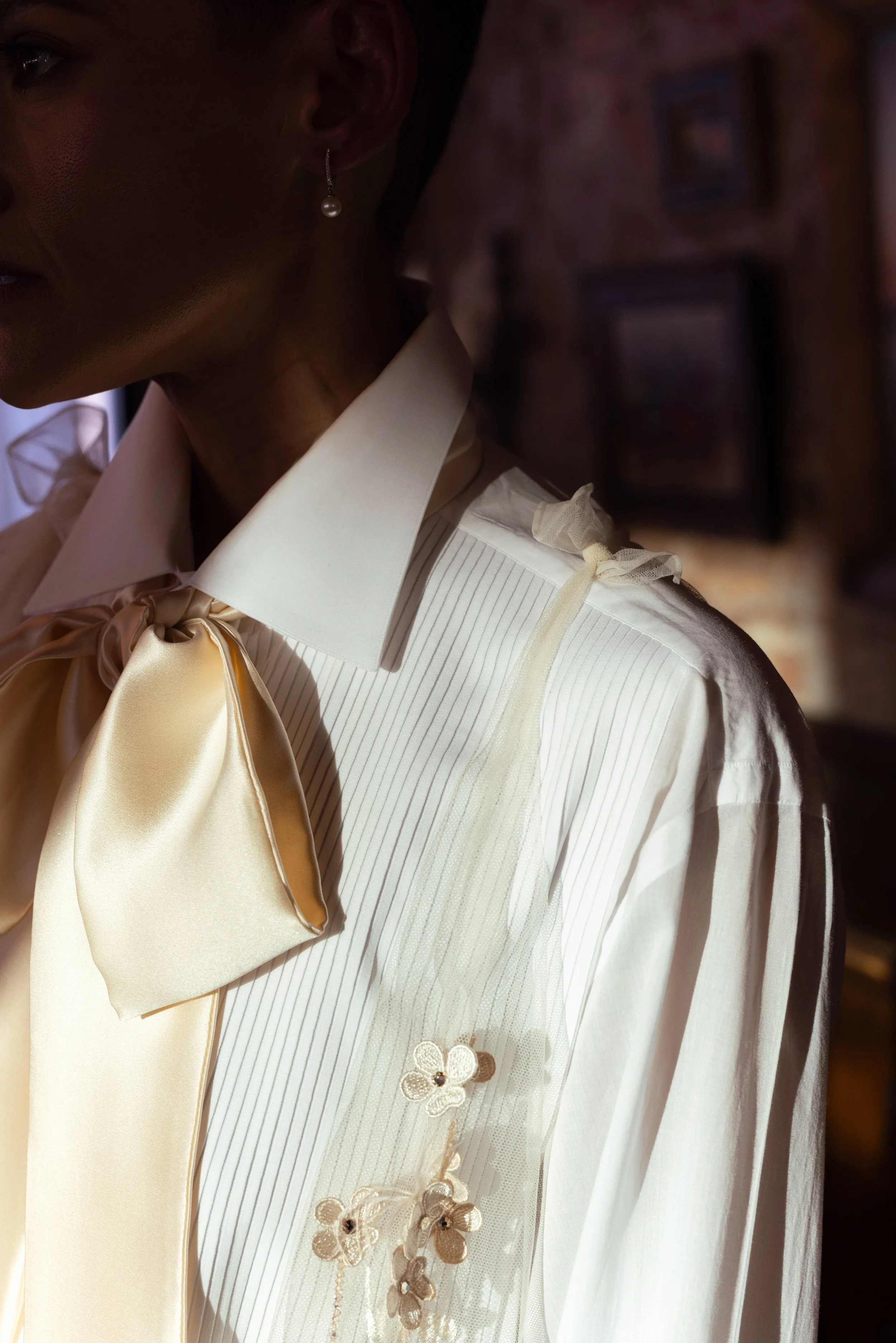 Close-up of a woman wearing a white Brioni tuxedo shirt with decorative floral embroidery, a large silk bow tie, and pearl and diamond earrings, in dim lighting with a blurred background.