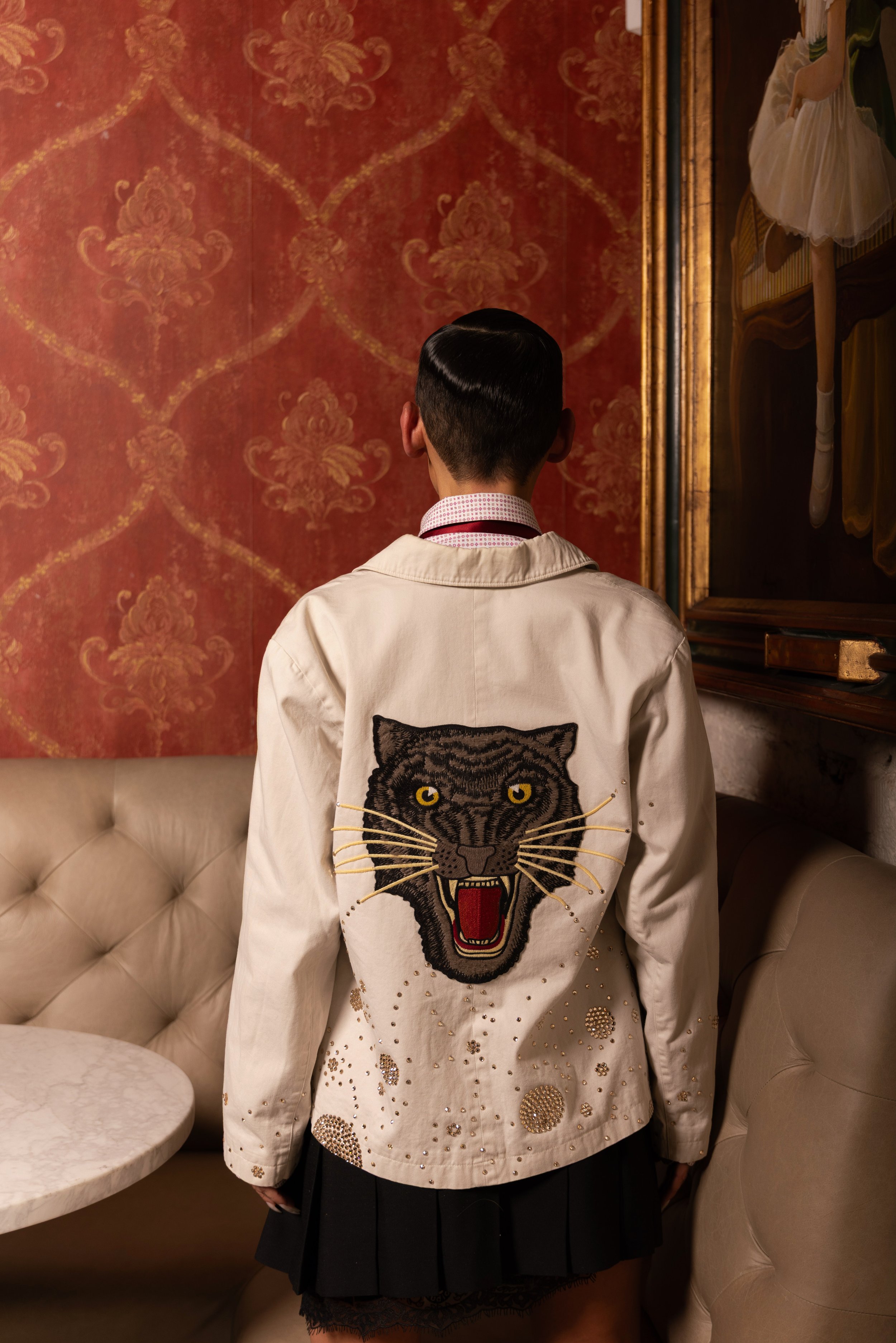 A person with short black hair is standing in front of a red patterned wall, wearing a cream-colored jacket with an embroidered fierce black panther face on the back, surrounded by gold embellishments.