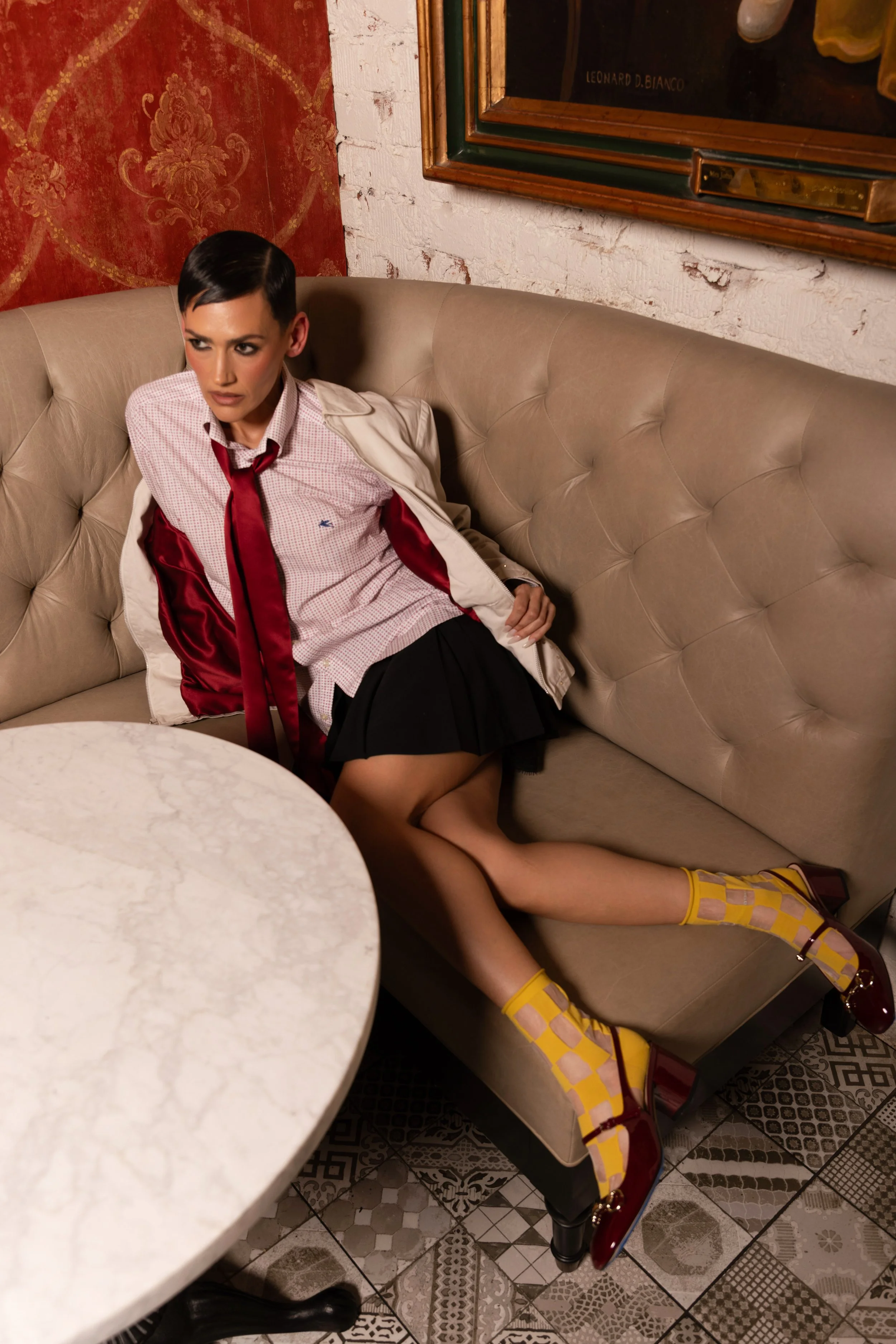 Young woman in a schoolgirl uniform sitting on a beige tufted sofa, wearing a red tie, checkered shirt, black skirt, and yellow checkered socks with burgundy high heels. She is leaning back with one leg extended and looking to the side.