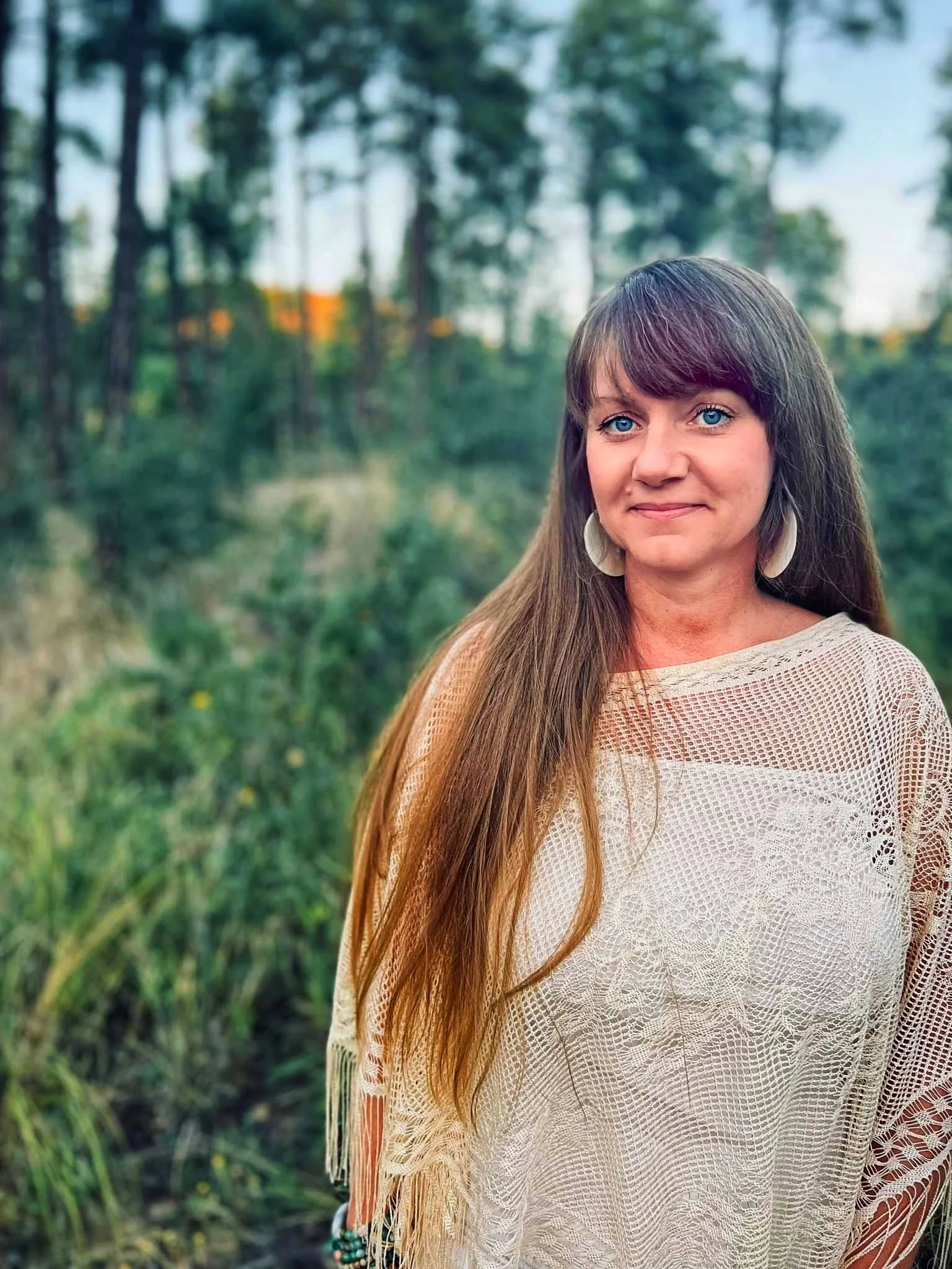 A woman with long brown hair and blue eyes standing outdoors in a green, wooded area, wearing a beige crocheted top with fringes.