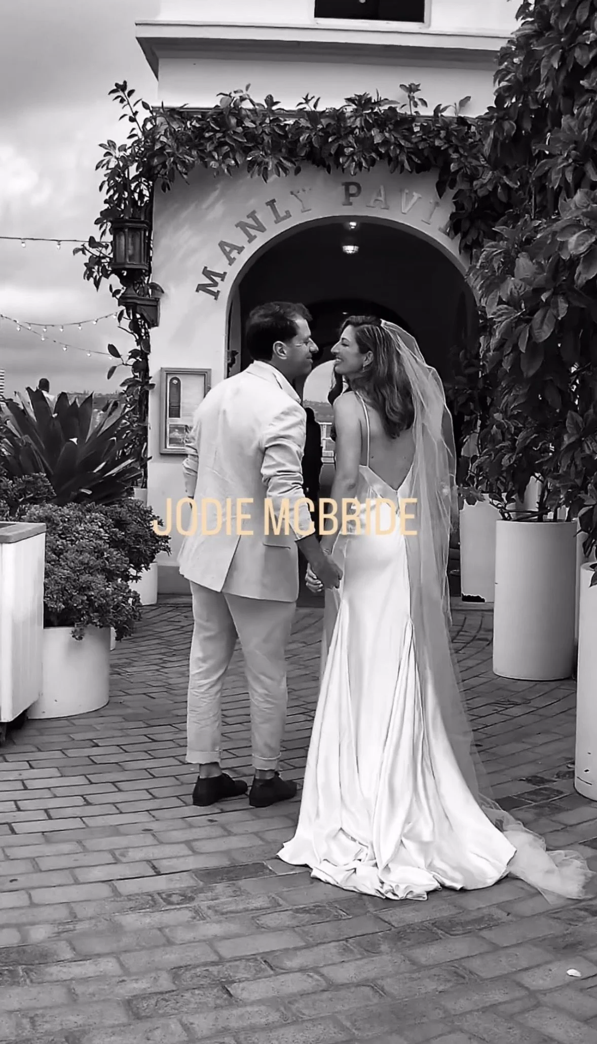 A black and white photo of a bride and groom holding hands and about to kiss, standing outside a building with the words 'MANLY PAVILION' above the archway. The groom wears a light-colored suit and the bride wears a long, elegant wedding gown with a veil. There are potted plants and string lights in the background.