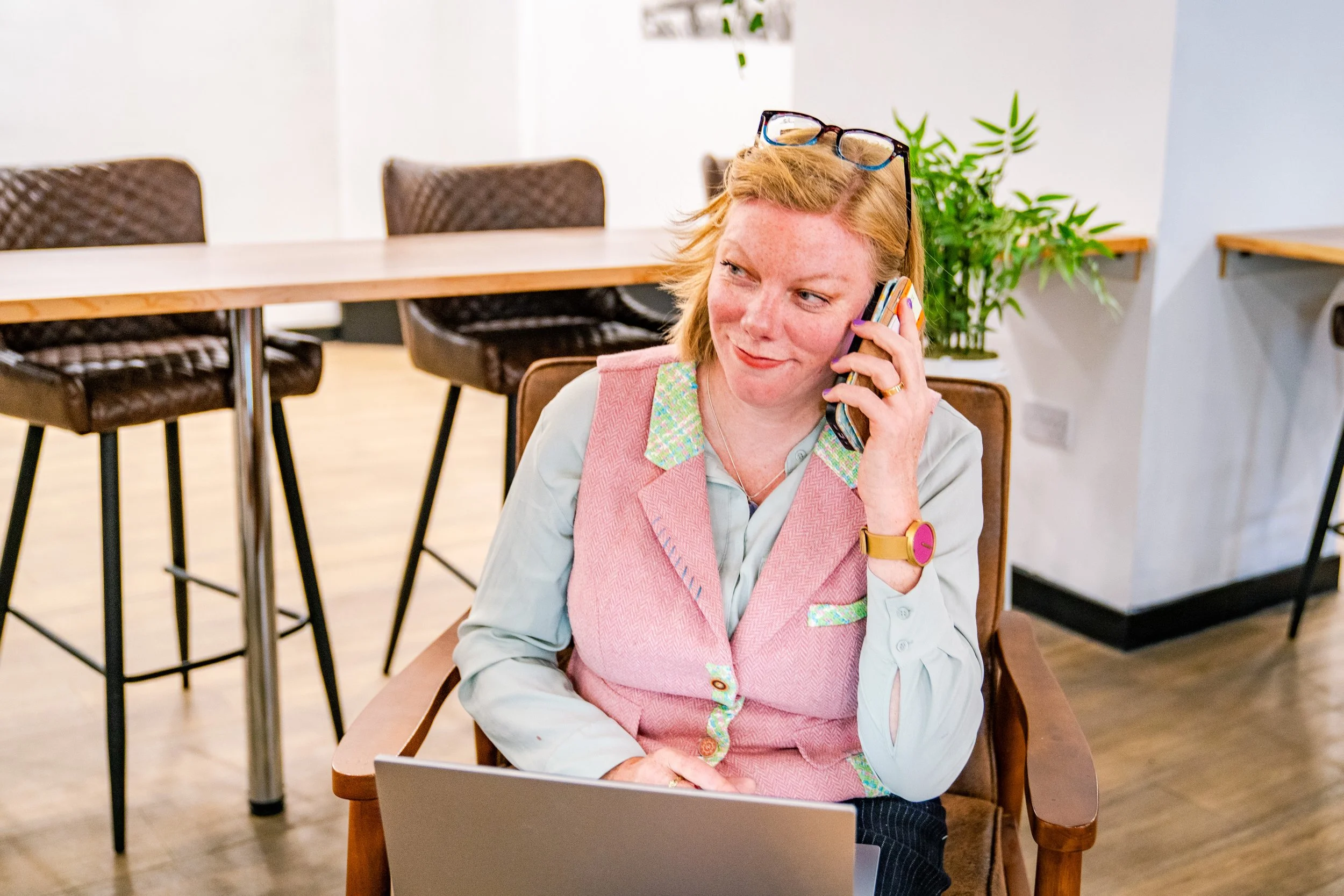 A woman with red hair, glasses on her head, wearing a pink vest with green accents, talking on a cell phone, sitting in a chair with a laptop in front of her. She is in a bright room with a wooden floor and a counter with barstools in the background. There is a green plant behind her.