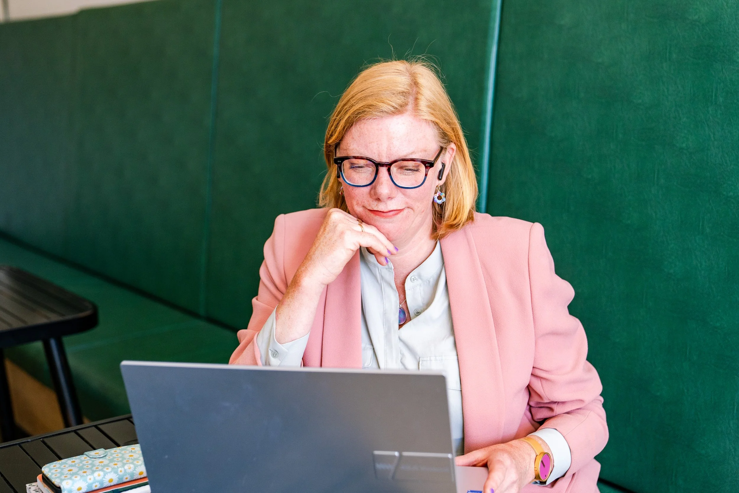 A woman with red hair and glasses, wearing a pink blazer and white shirt, sitting at a table and working on a laptop.