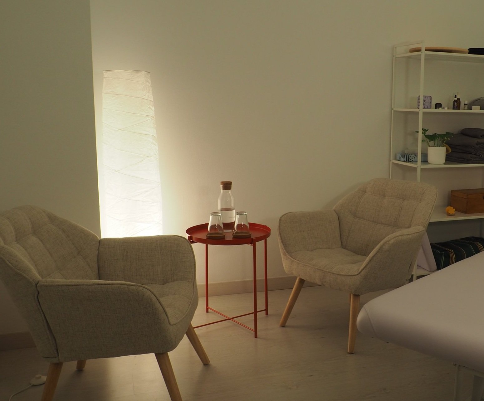 Alquiler de Sala con dos sillones beige de madera, mesa roja con un jarro y dos vasos, estantería blanca con objetos y plantas, lámpara de pie encendida.
