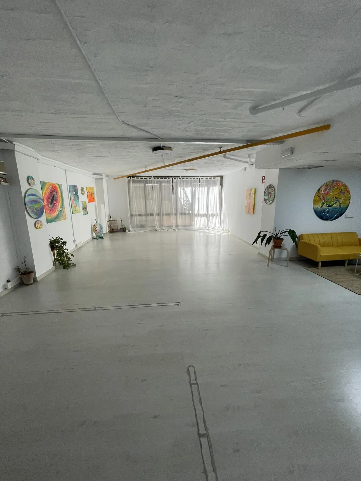 Alquiler Espacio amplio y luminoso con paredes blancas y decoraciones coloridas, incluyendo pinturas en la pared, plantas en macetas, y un sillón amarillo en un rincón.
