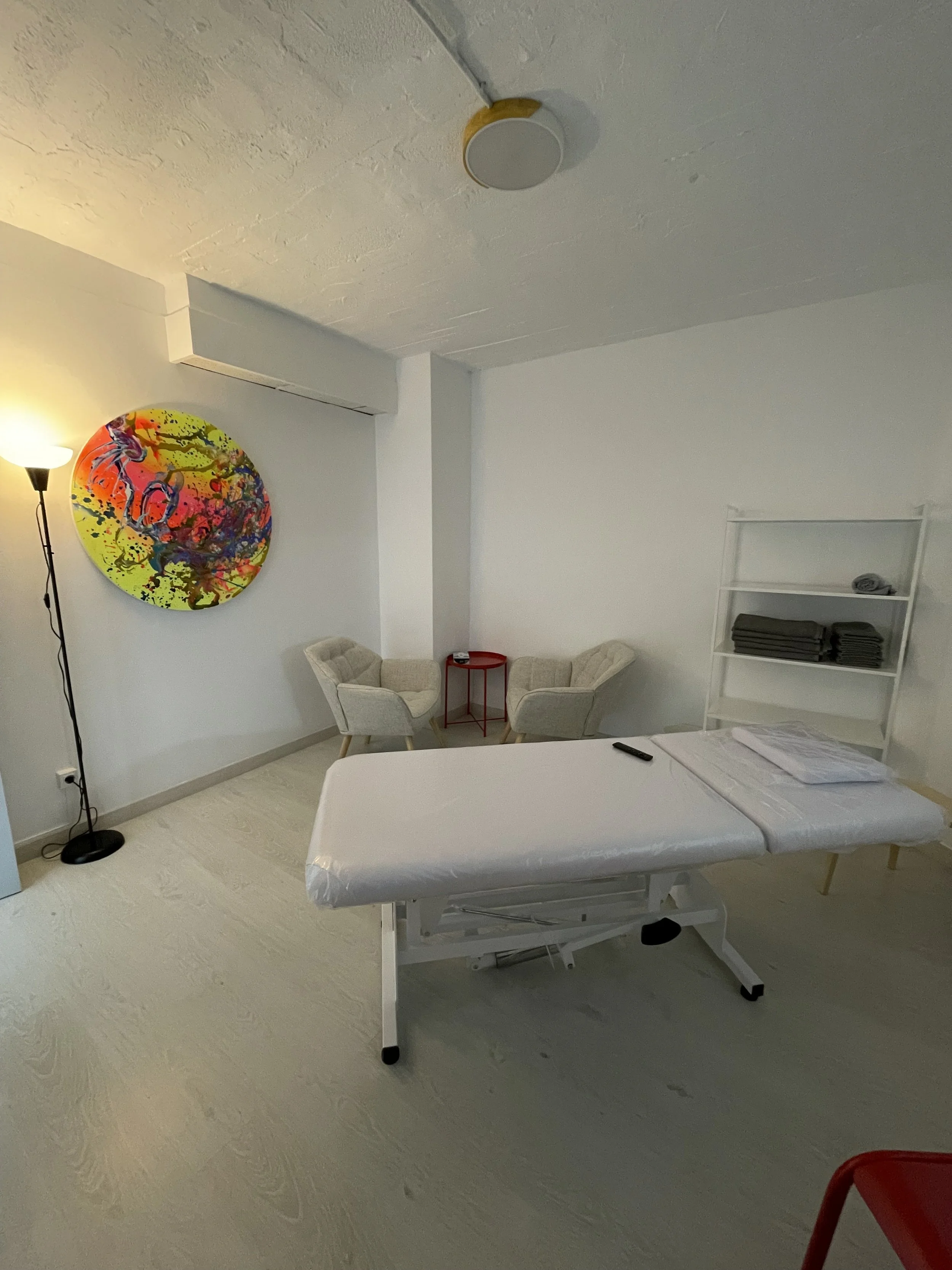 Alquiler Una sala con una camilla de masaje, una lámpara de pie, una pared con una obra de arte colorida, una pequeña mesa roja, una silla y un estante con toallas.