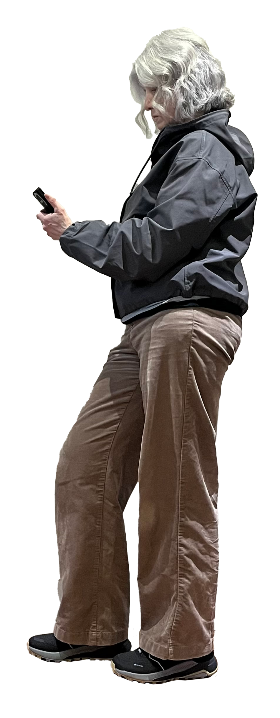 Older woman with gray hair looking at her phone, dressed in a gray jacket, brown pants, and black shoes, standing against a black background.
