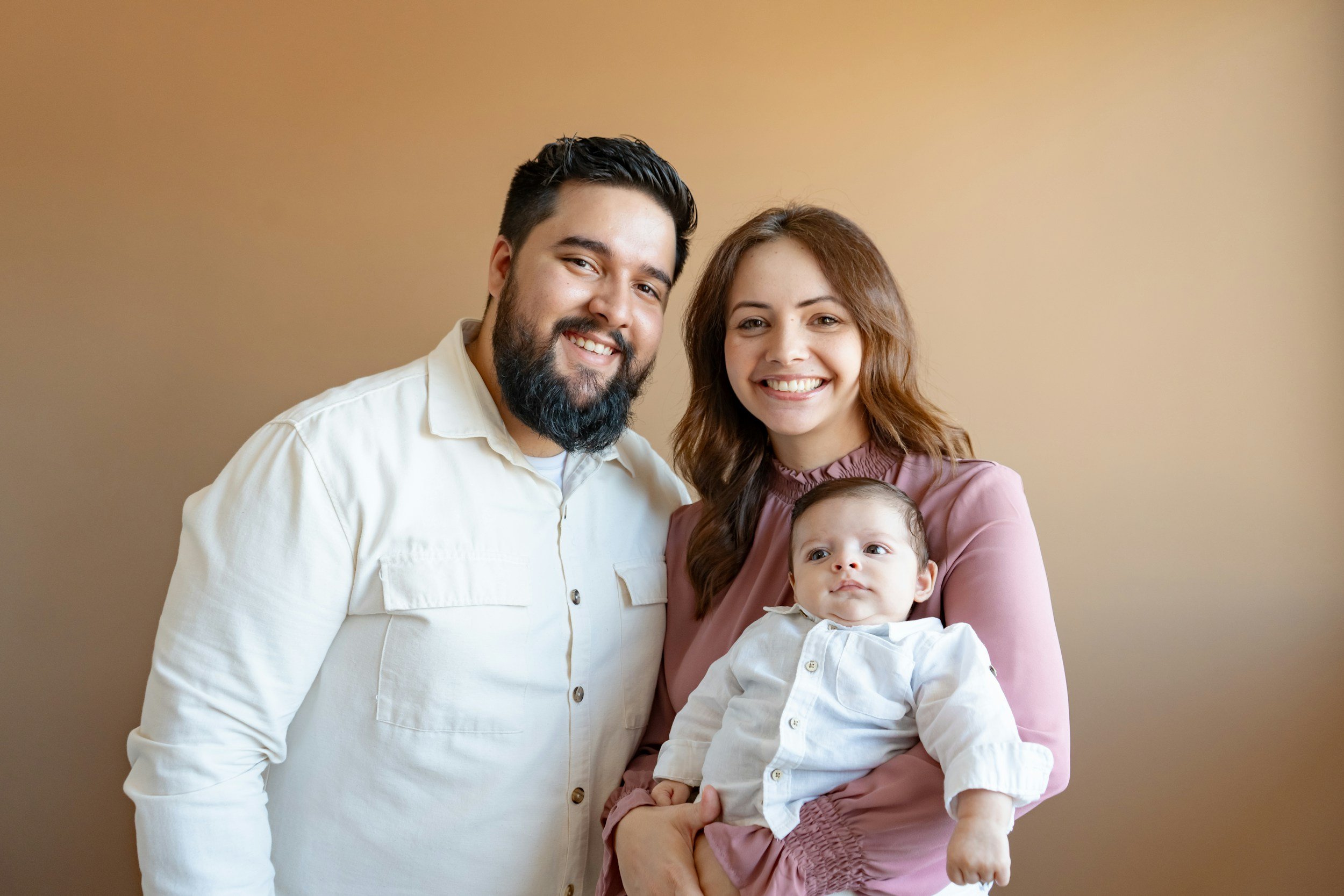 Father and mother stand in front of plain background. Mother is holding a 8 month old baby. Family are from middle-eastern  background.