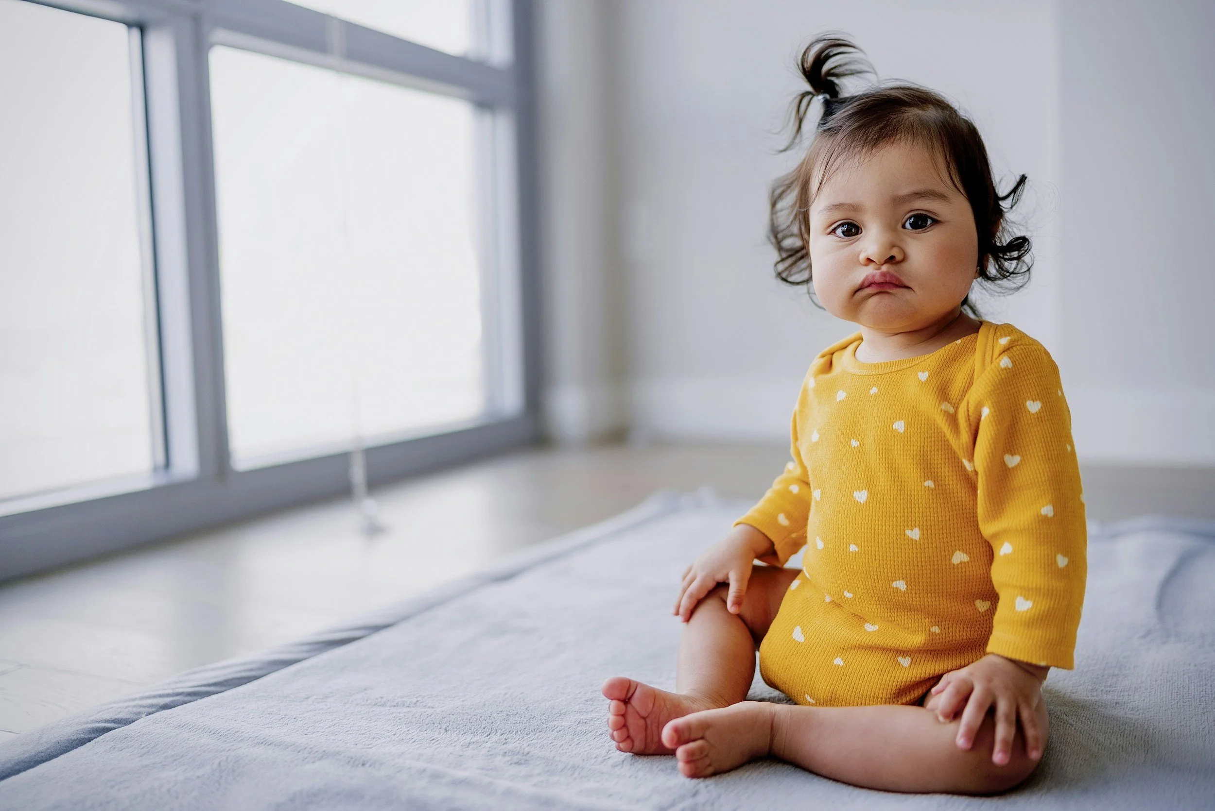 Nine month old baby girl sits up on mat in front of a window.