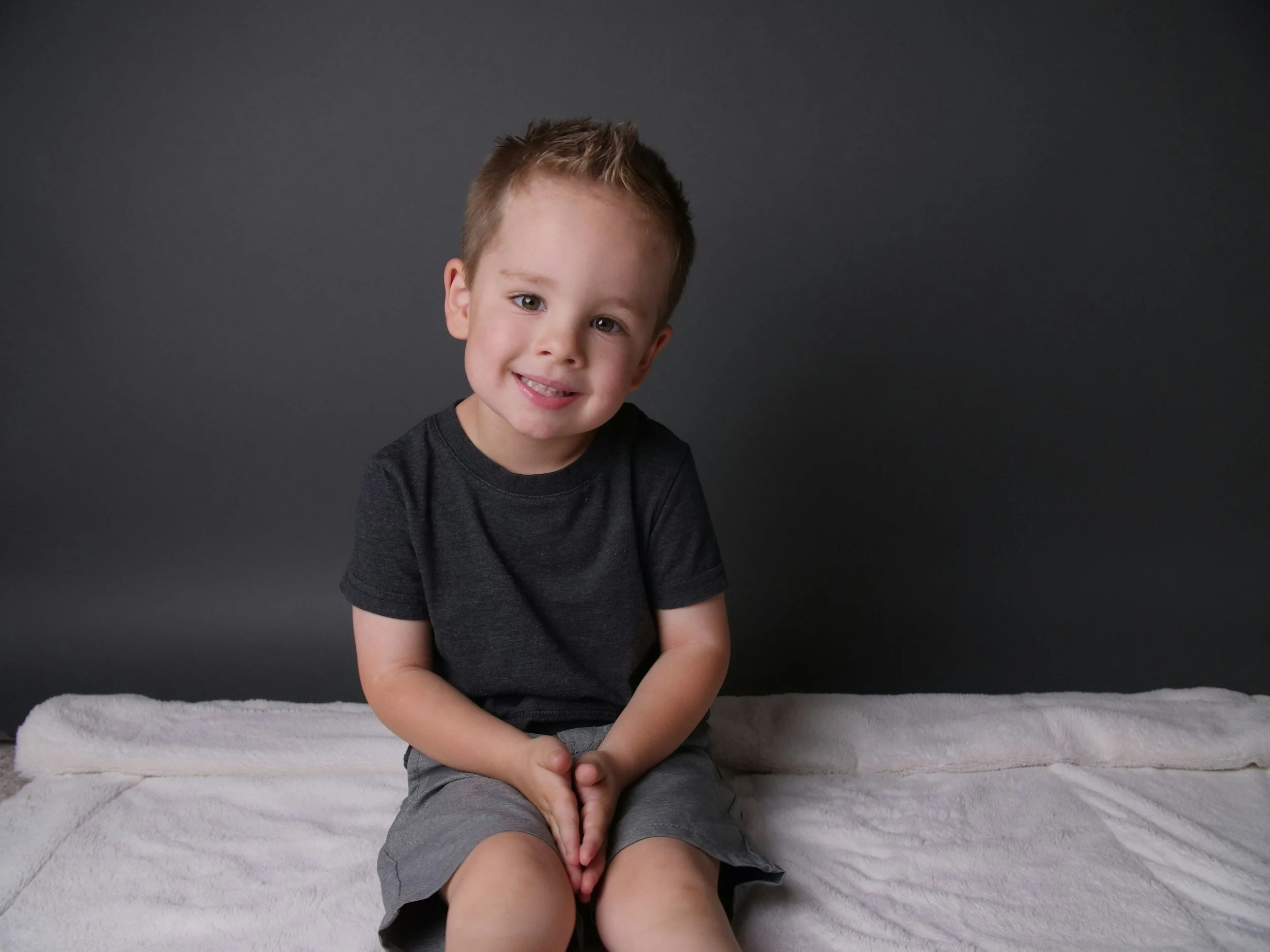 Three year old boy sits on a bed in front of a dark grey wall.
