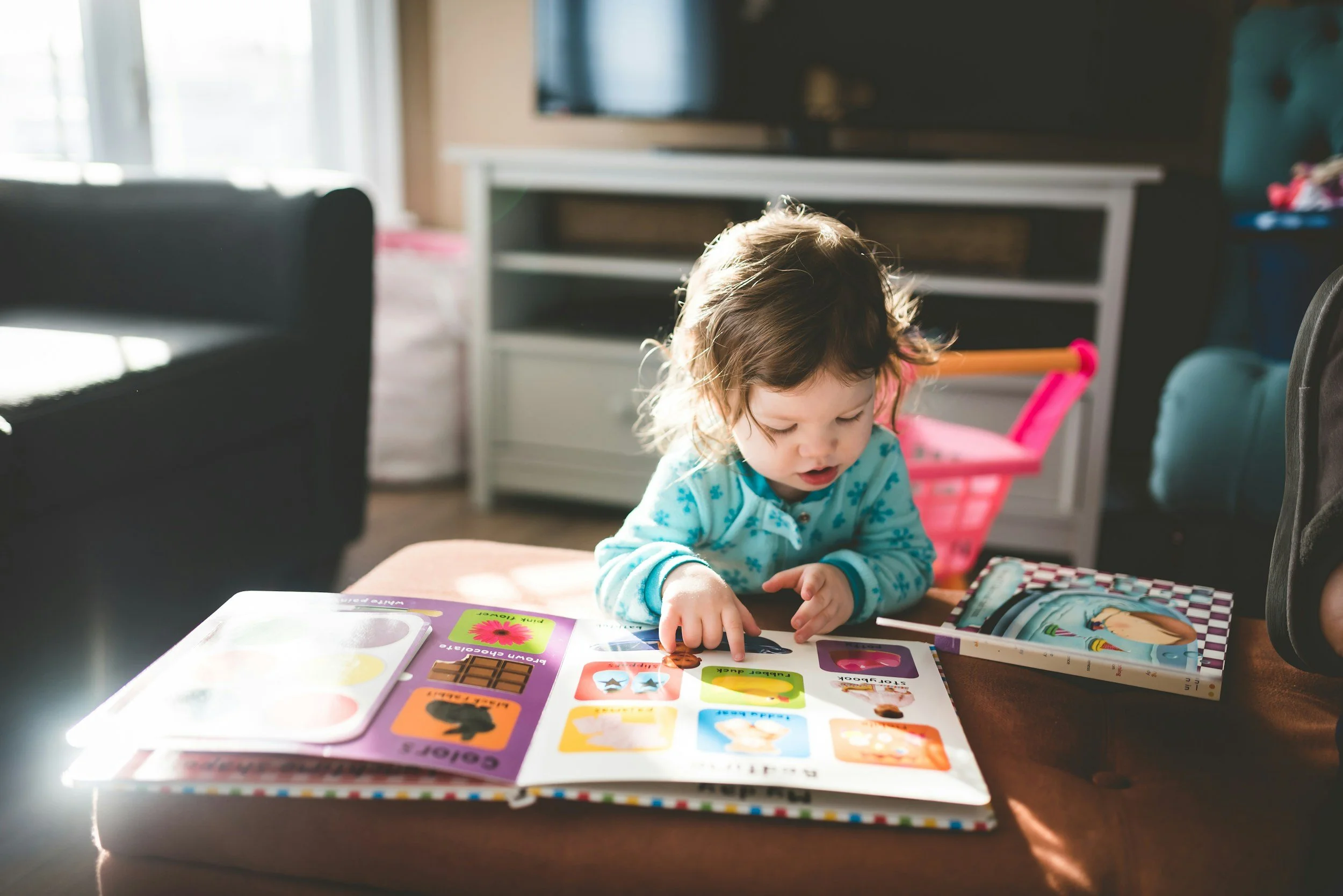 A toddler stands at a brown ottoman and points to pictures in a children's picture book.