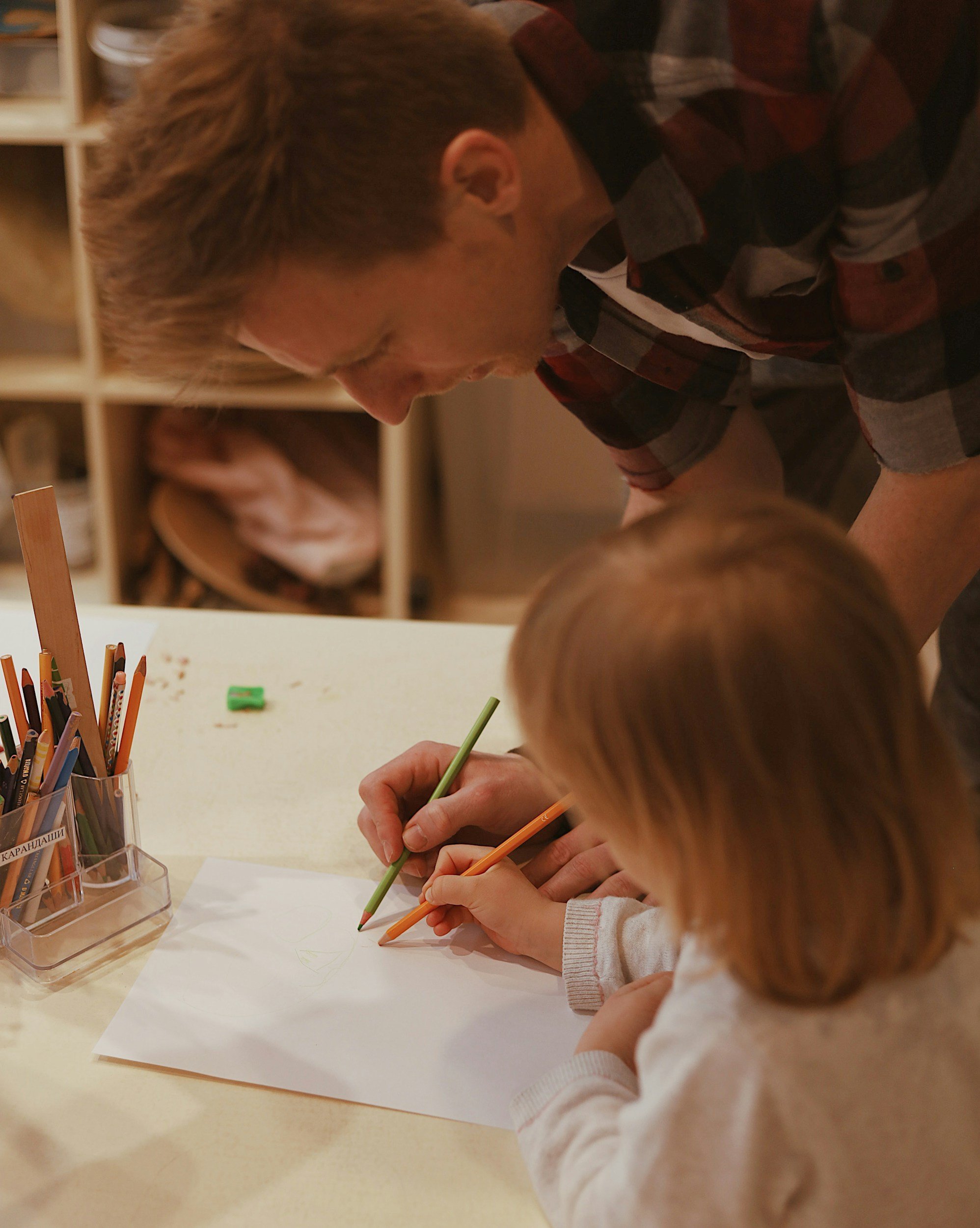 A dad draws with his child. Both the dad and child hold pencils and look towards the paper.