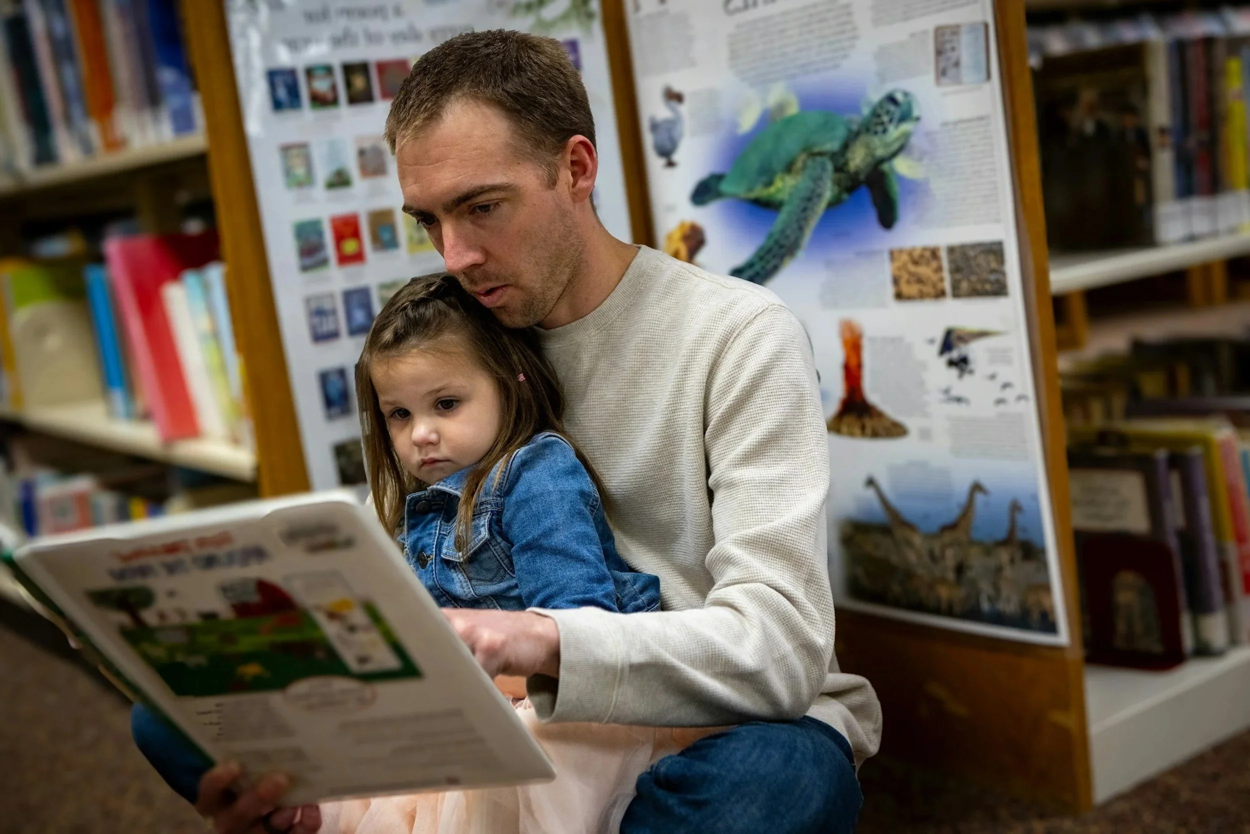 Girl sits on father's lap in a library, while he reads her a book.