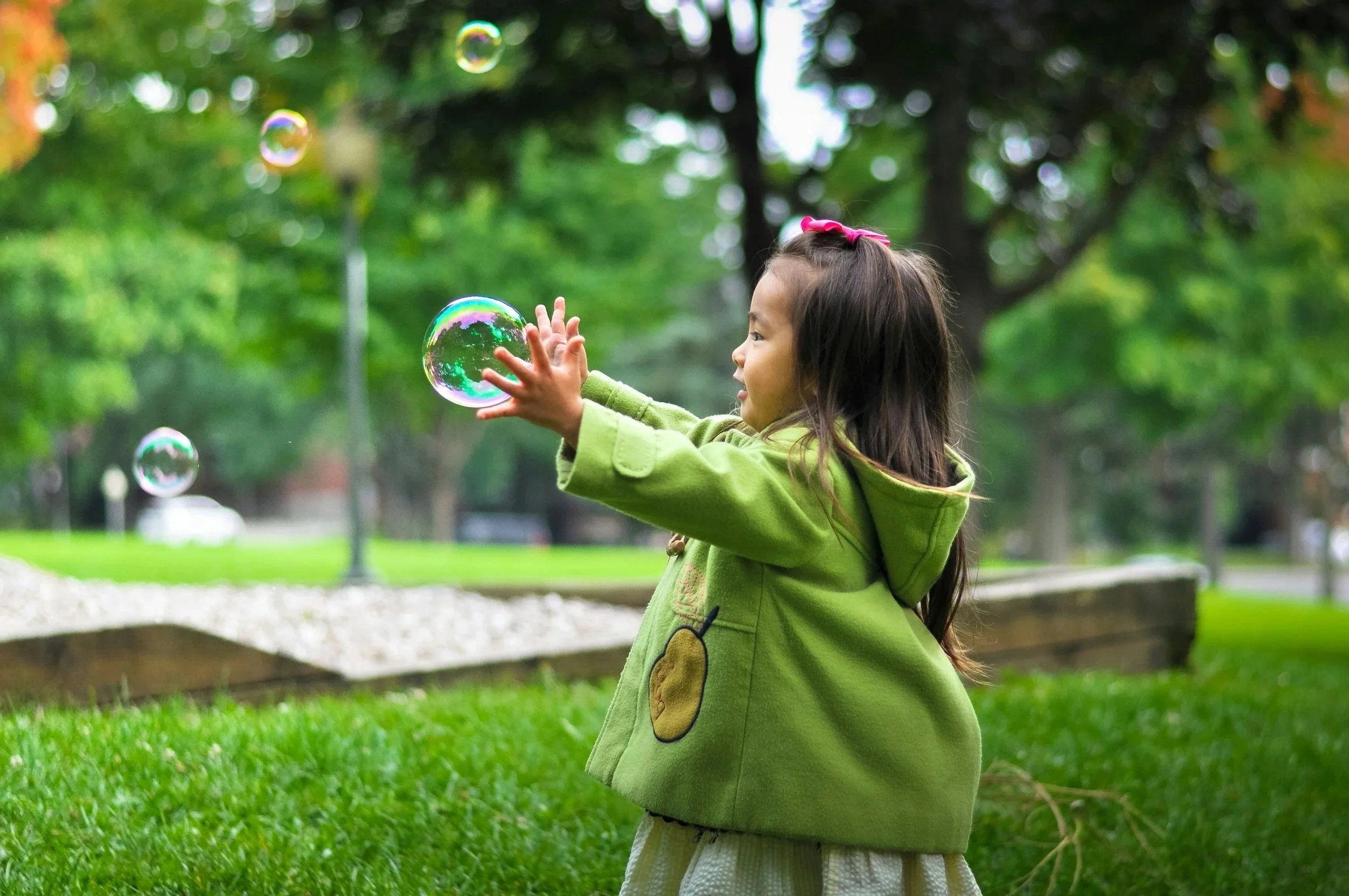 Young girl wearing a green jacket is playing with bubbles in a park.
