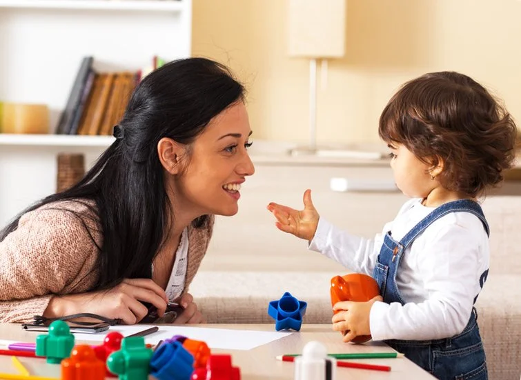 Mother plays with child at coffee table. There are a range of shape blocks and coloured pens on the table.