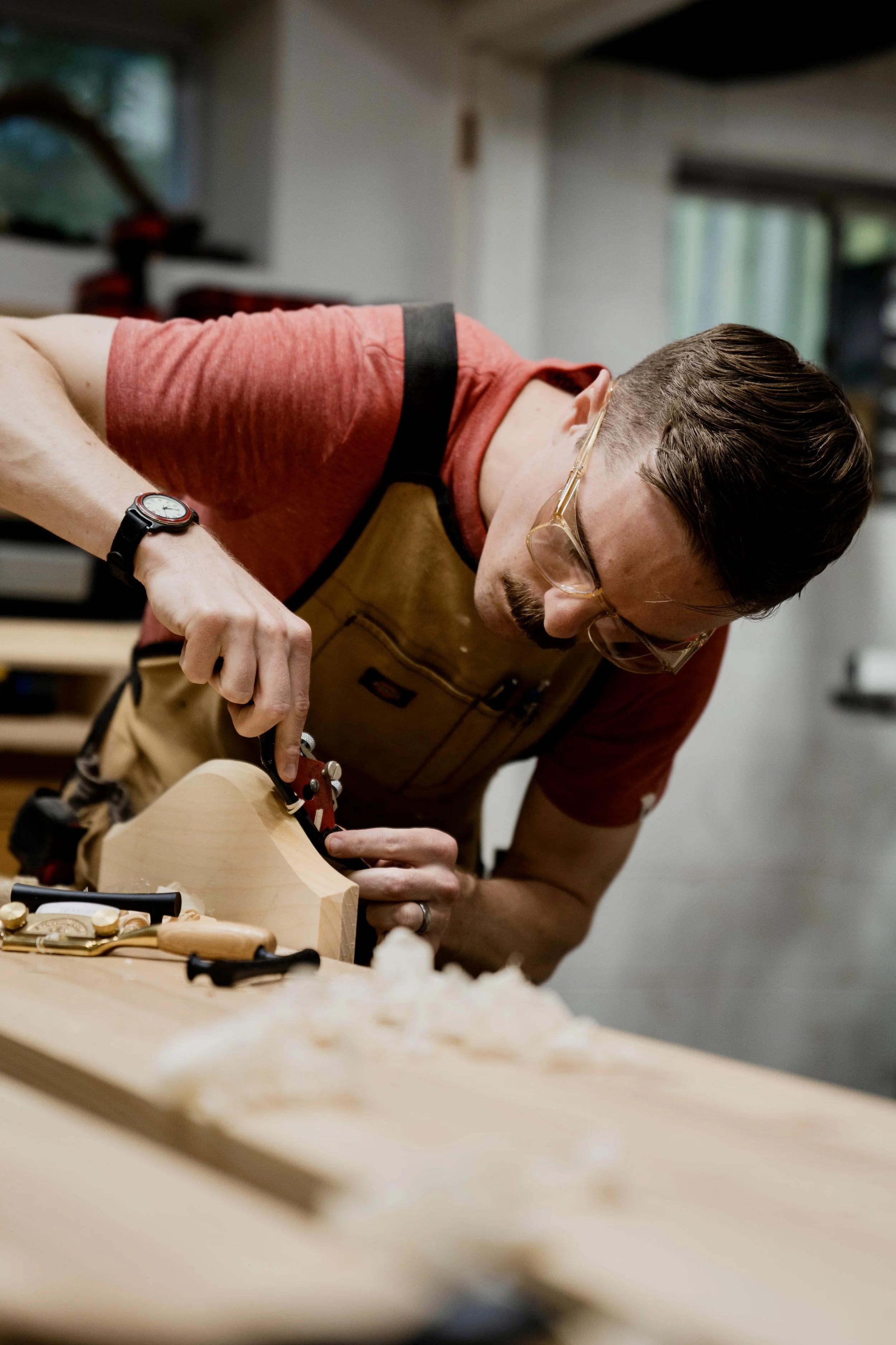 A man wearing glasses, a red shirt, and a brown apron is woodworking, using a tool on a piece of wood, in a workshop.