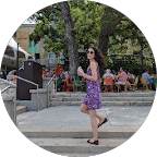 A woman in a purple floral dress walking outdoors in a park or city square.