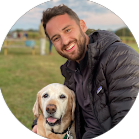 A smiling man with a beard outdoors holding a Labrador Retriever dog.