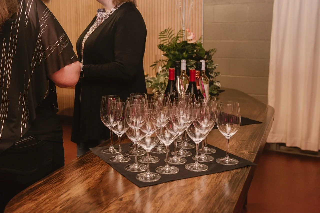 Empty wine glasses arranged on a black mat on a wooden table, with bottles of wine and a plant in the background, at an indoor event.