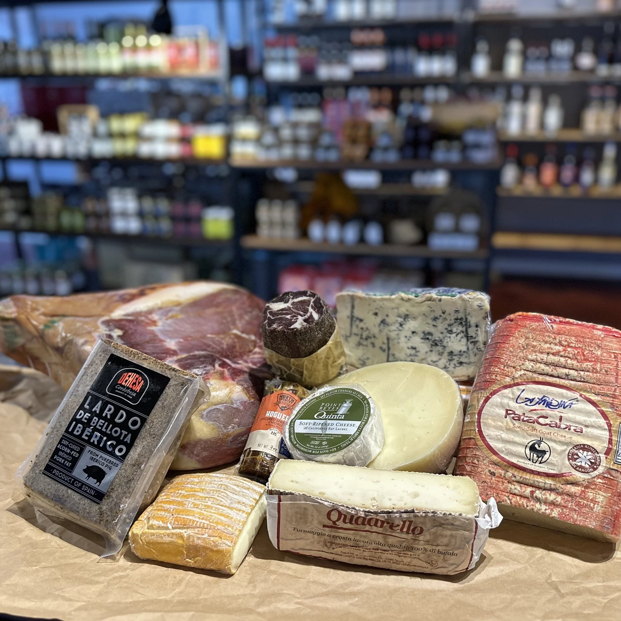 Various cheeses, a piece of Iberico ham, and a bundle of black truffles on a wooden surface in a store.